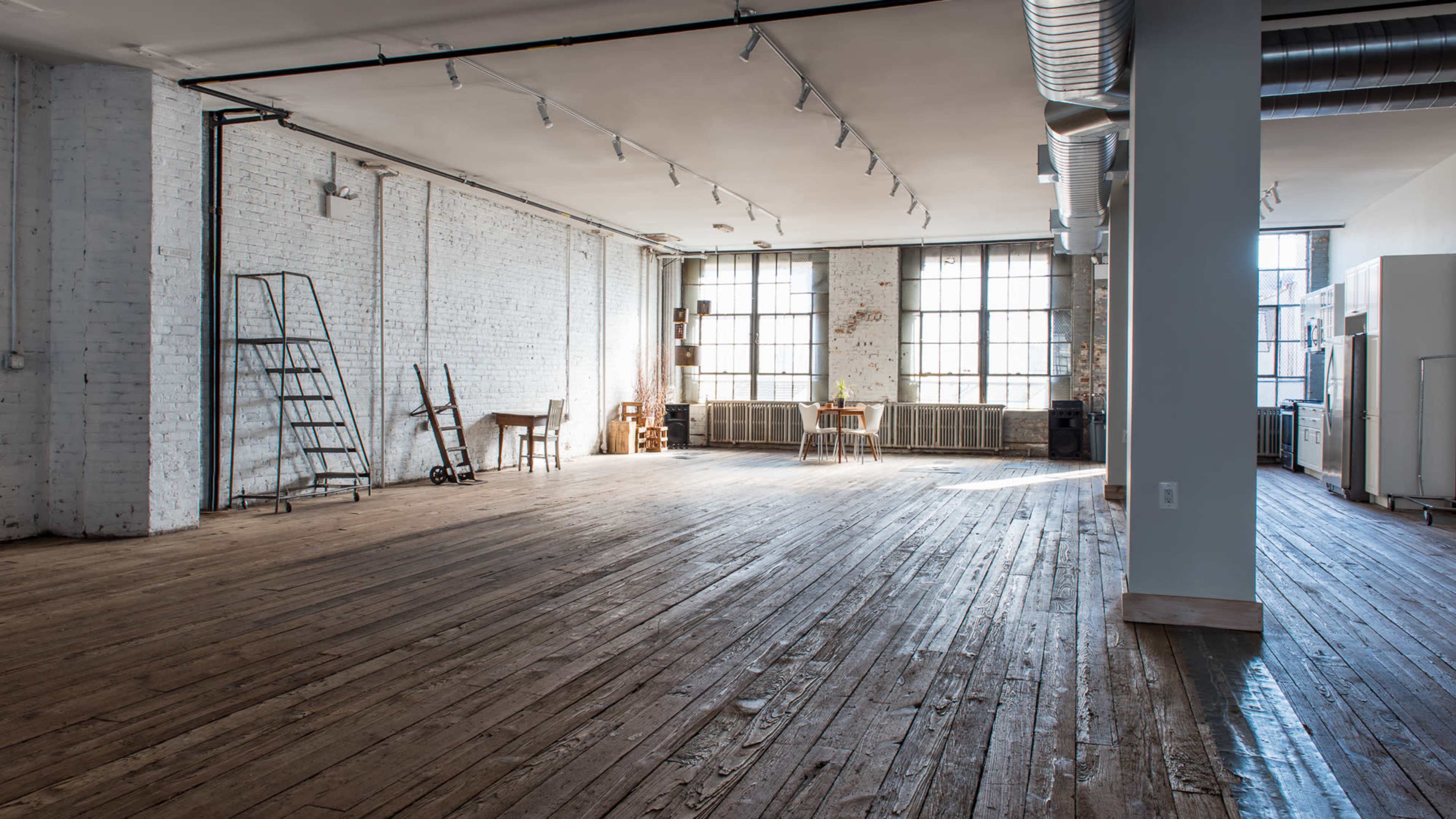A spacious, empty loft with wooden floors, exposed brick walls, large windows, and minimal furnishings.