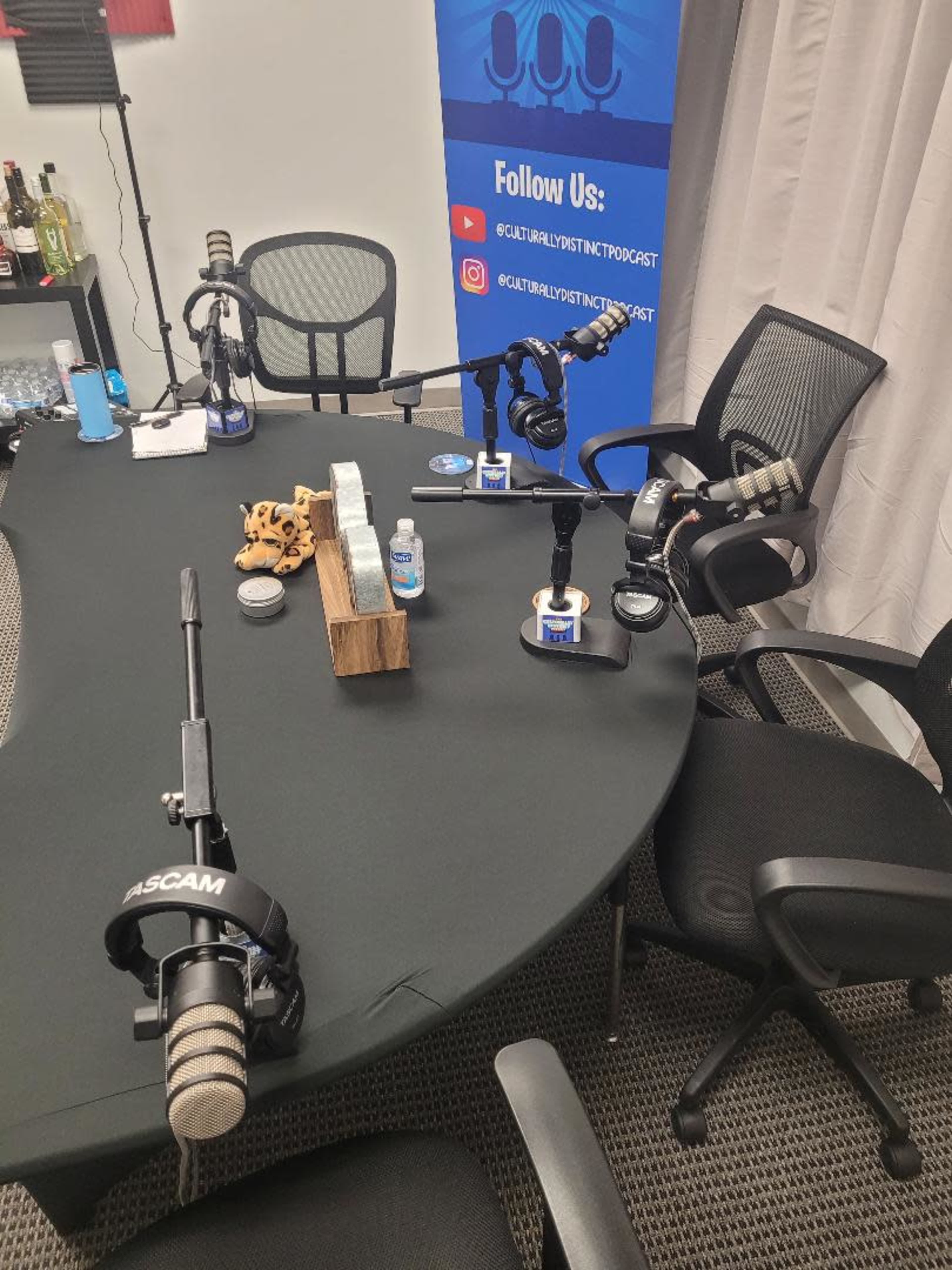 The image shows a podcast recording setup with four microphones, chairs, and a central wooden display on a round black table.