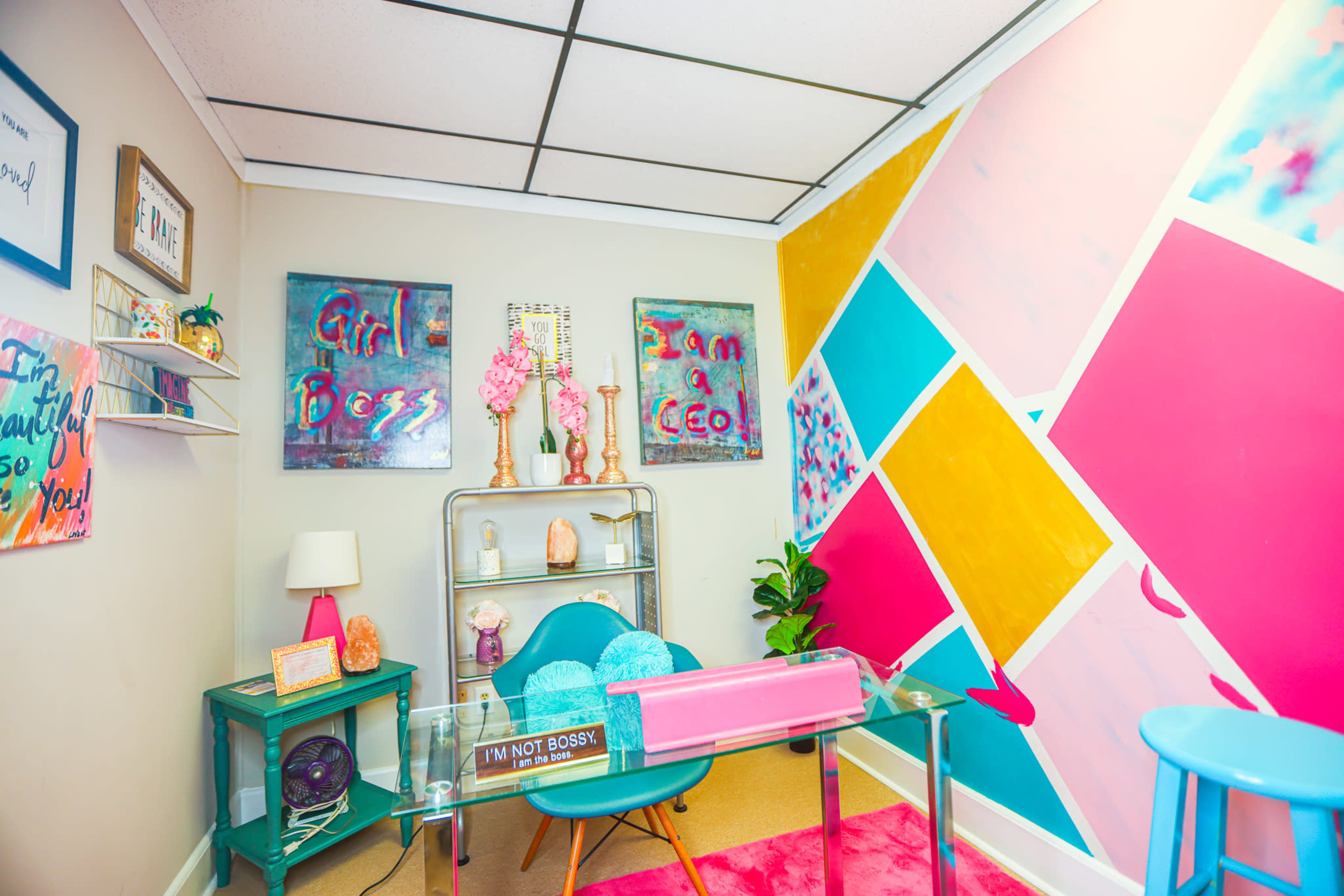 A small office space featuring a brightly colored accent wall, a glass desk with a pink and clear design, and decorative elements like artworks and plants.