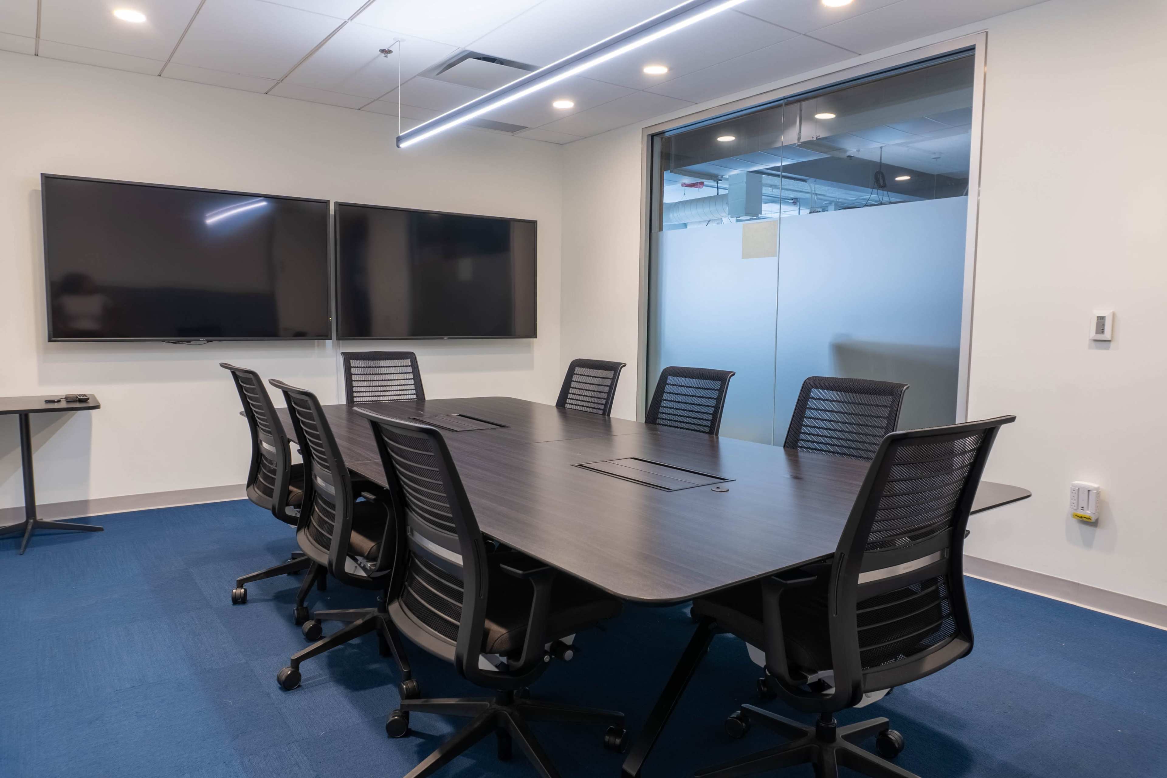 A modern conference room with a large dark table, eight rolling chairs, and two wall-mounted screens.