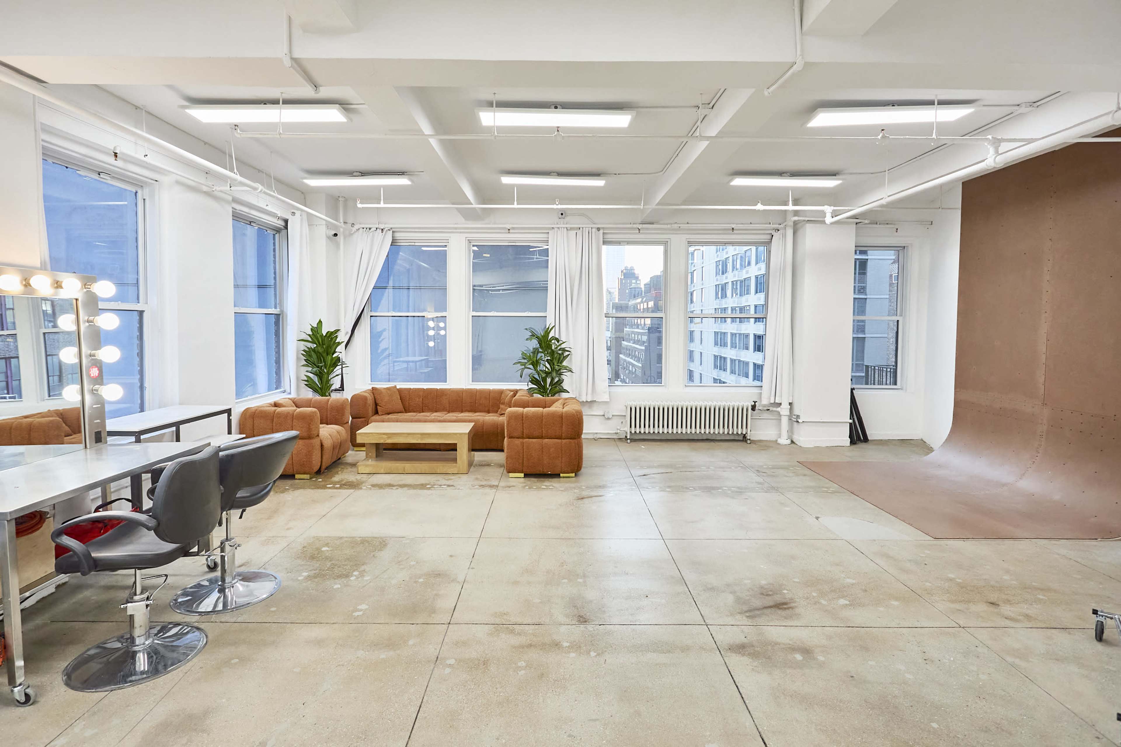 The scene depicts a bright, spacious studio with large windows, modern furnishings, and a minimalist design featuring a lounge area and a backdrop for photography.