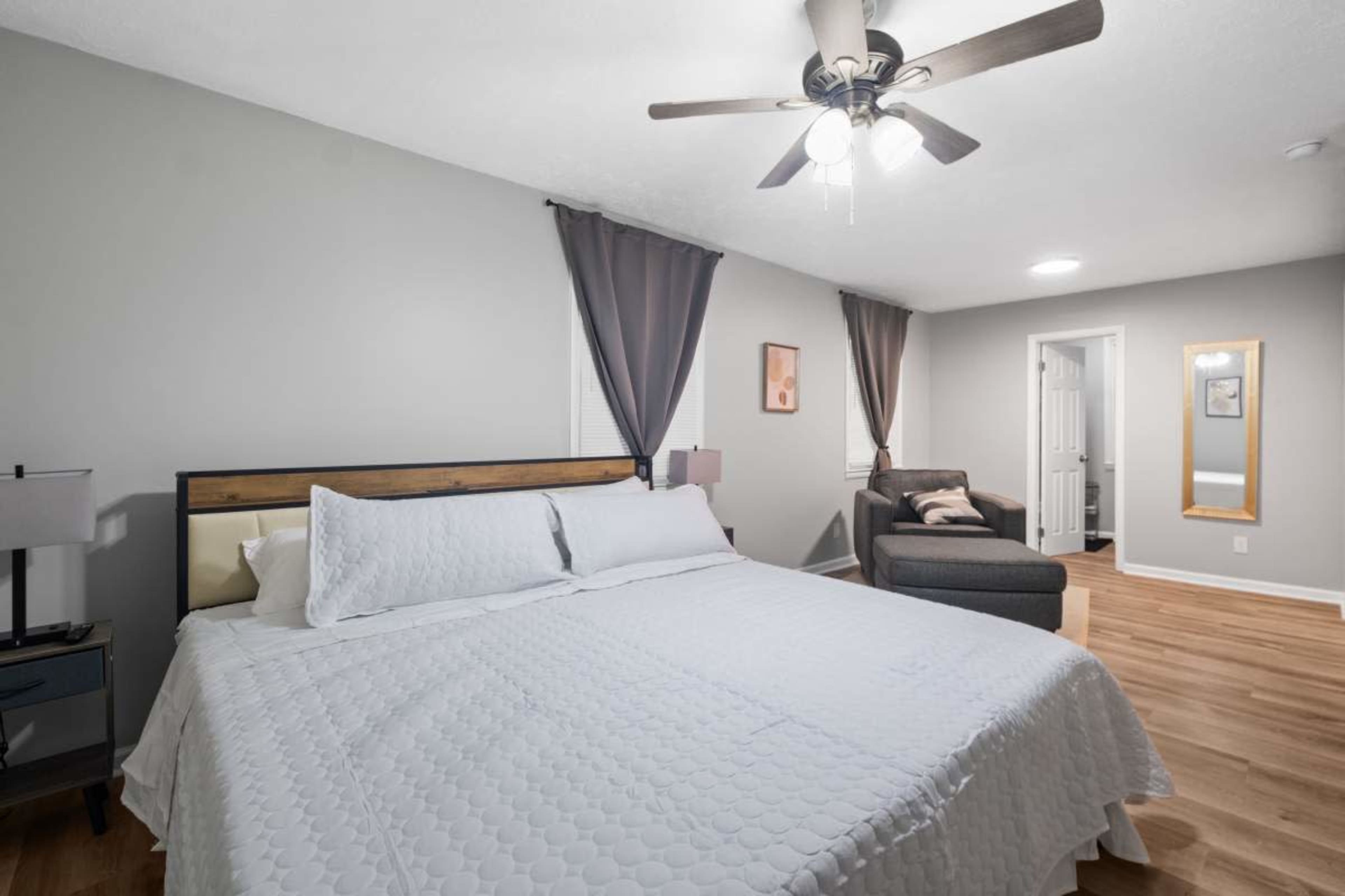 The image depicts a neatly arranged bedroom featuring a large bed with a light-colored quilt, a ceiling fan, and a seating area with a gray sofa.