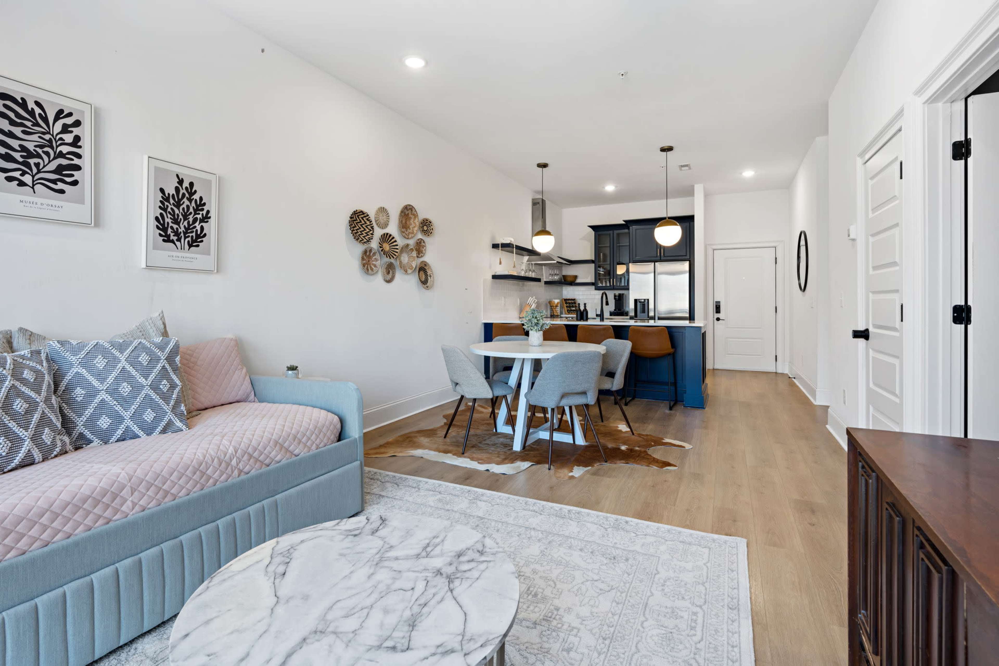 The image shows a modern living space featuring a pastel pink sofa, a circular marble coffee table, a dining area with light blue chairs, and a kitchen in the background.