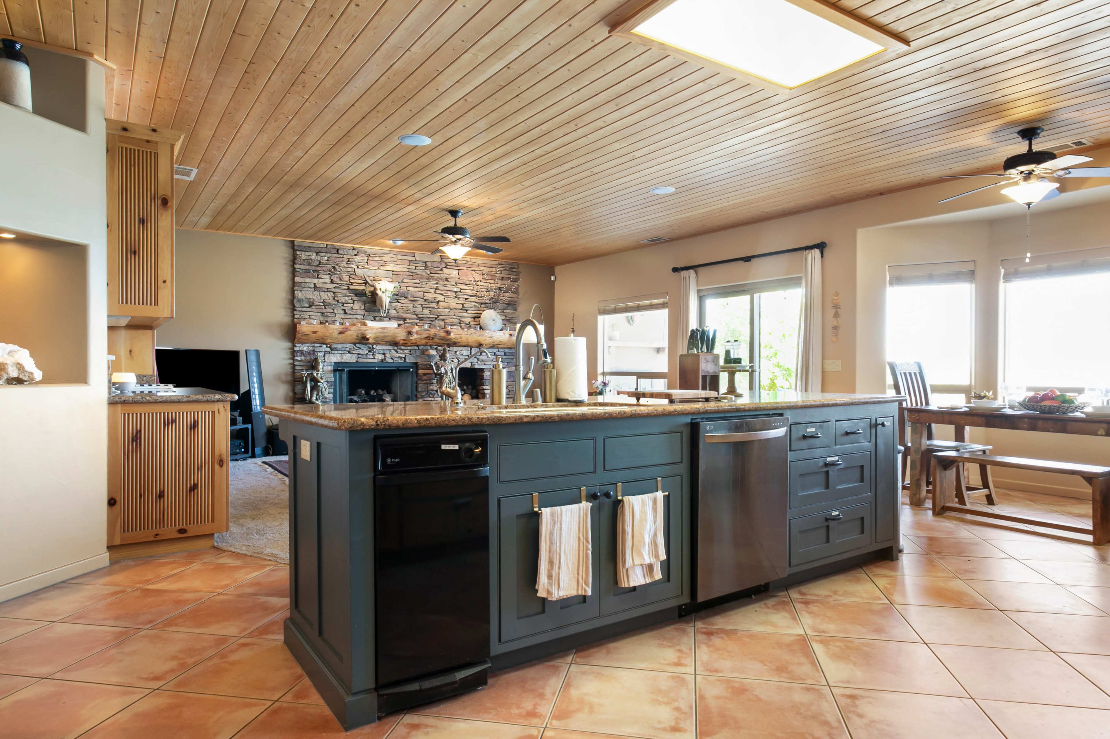 A modern kitchen features a central island with a dark countertop, stainless steel appliances, and wooden ceiling beams, adjacent to a living area with a stone fireplace and large windows.