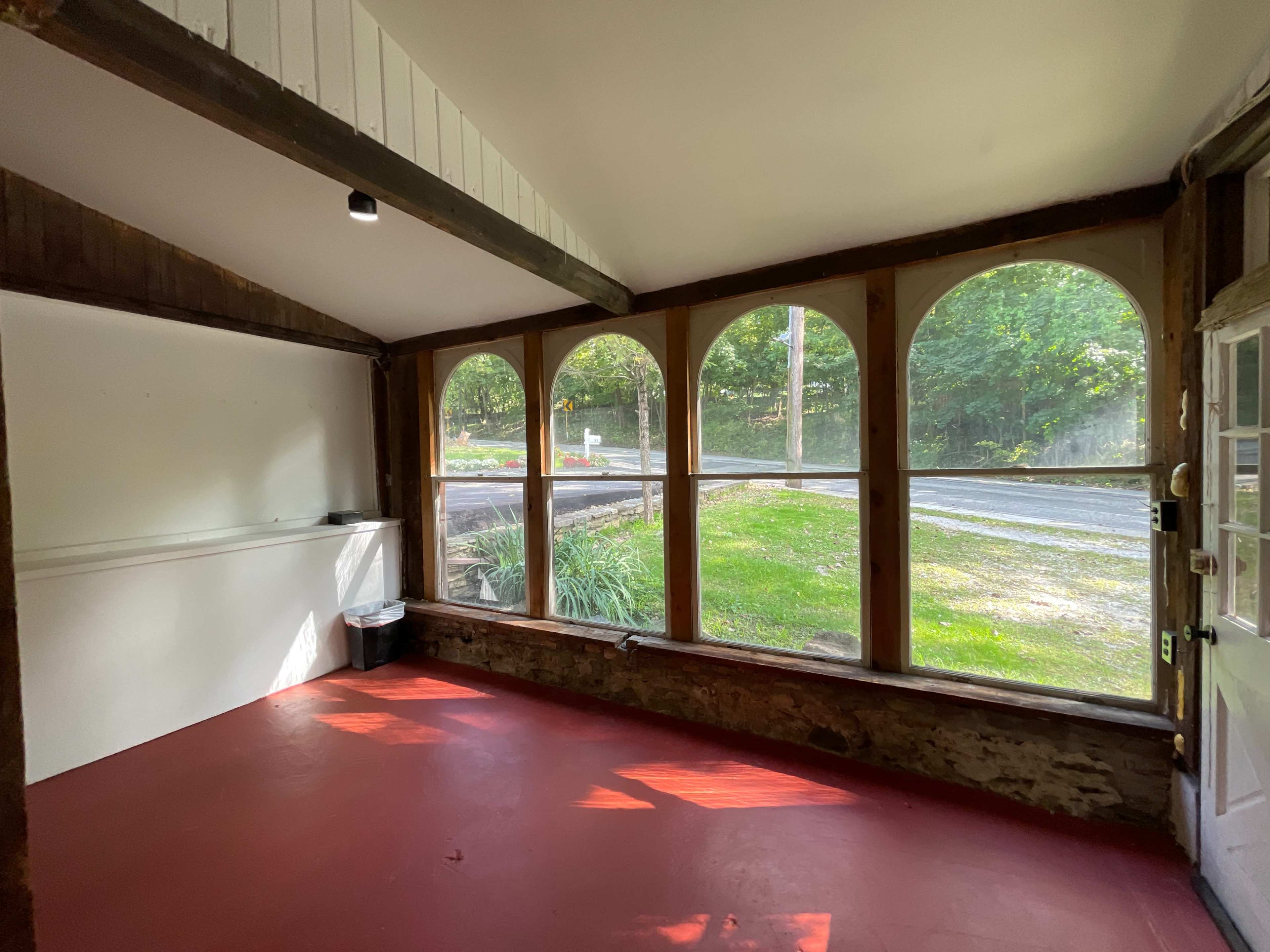 An empty, sunlit room with large arched windows overlooking a grassy area and a road outside.