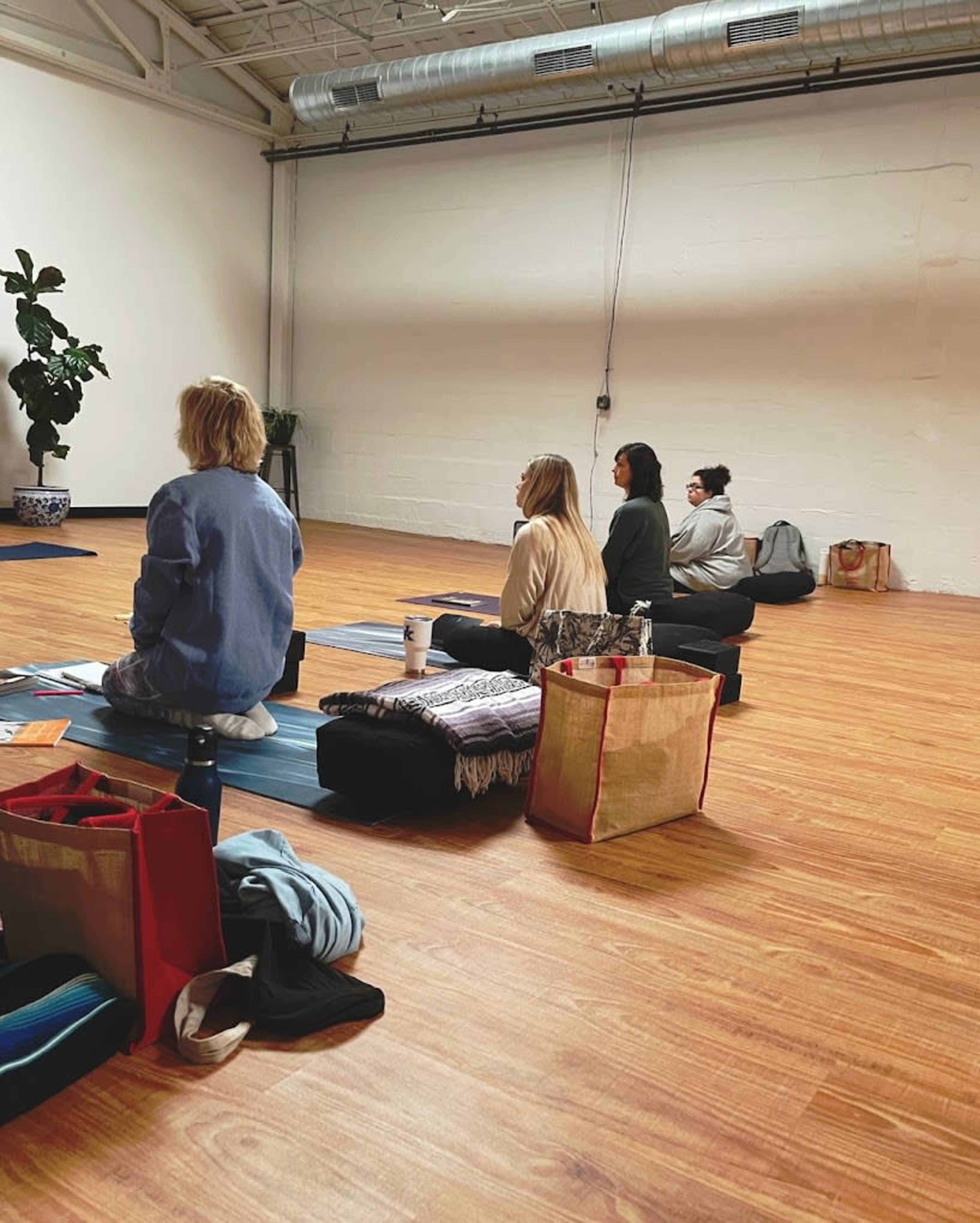 A group of individuals sit in a yoga studio, facing forward on mats with bags and personal items beside them.