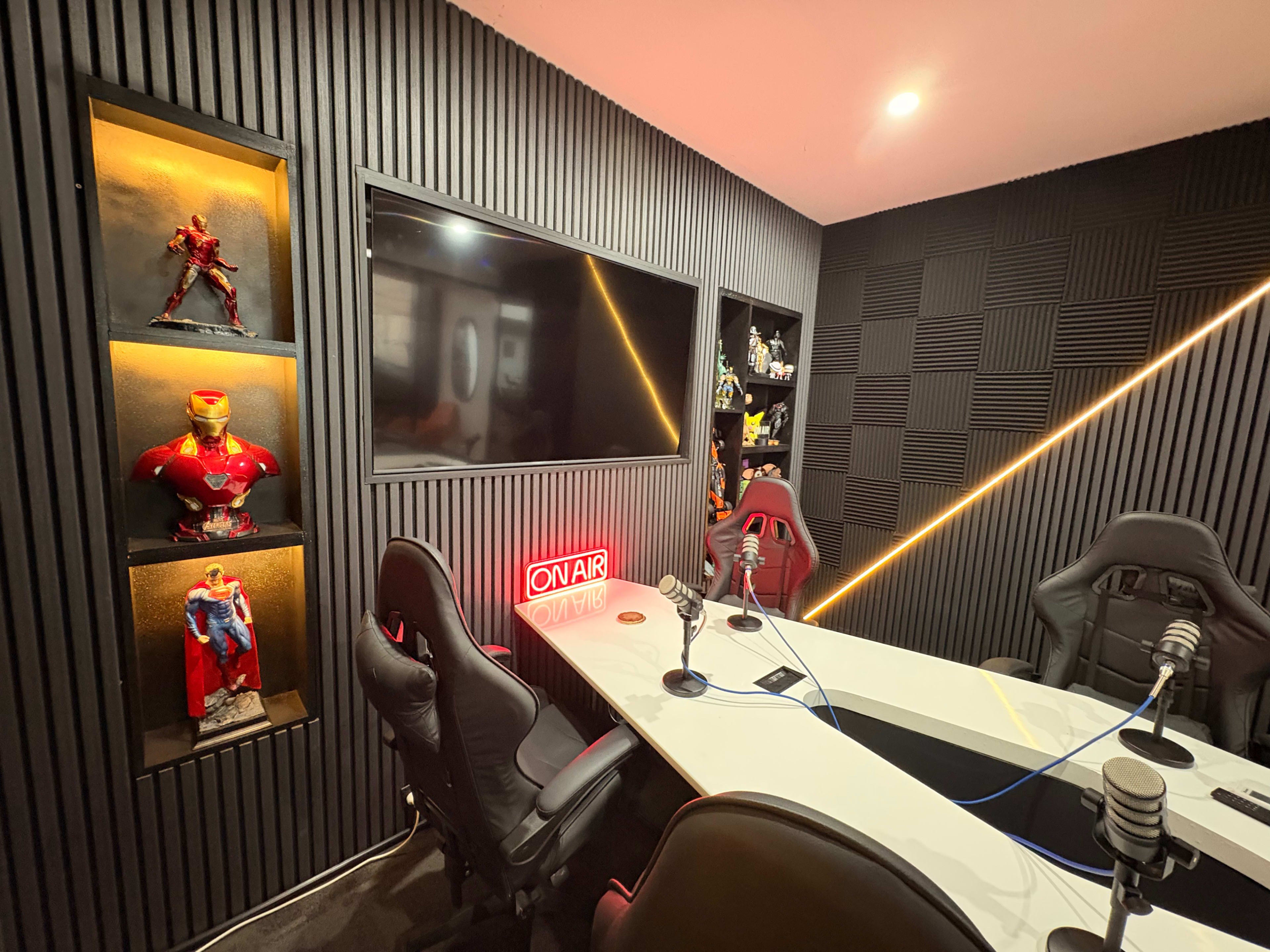 The image shows a modern recording studio featuring a long, white table surrounded by black and red gaming chairs, with superhero figurines displayed on wall shelves and a large screen mounted in front.