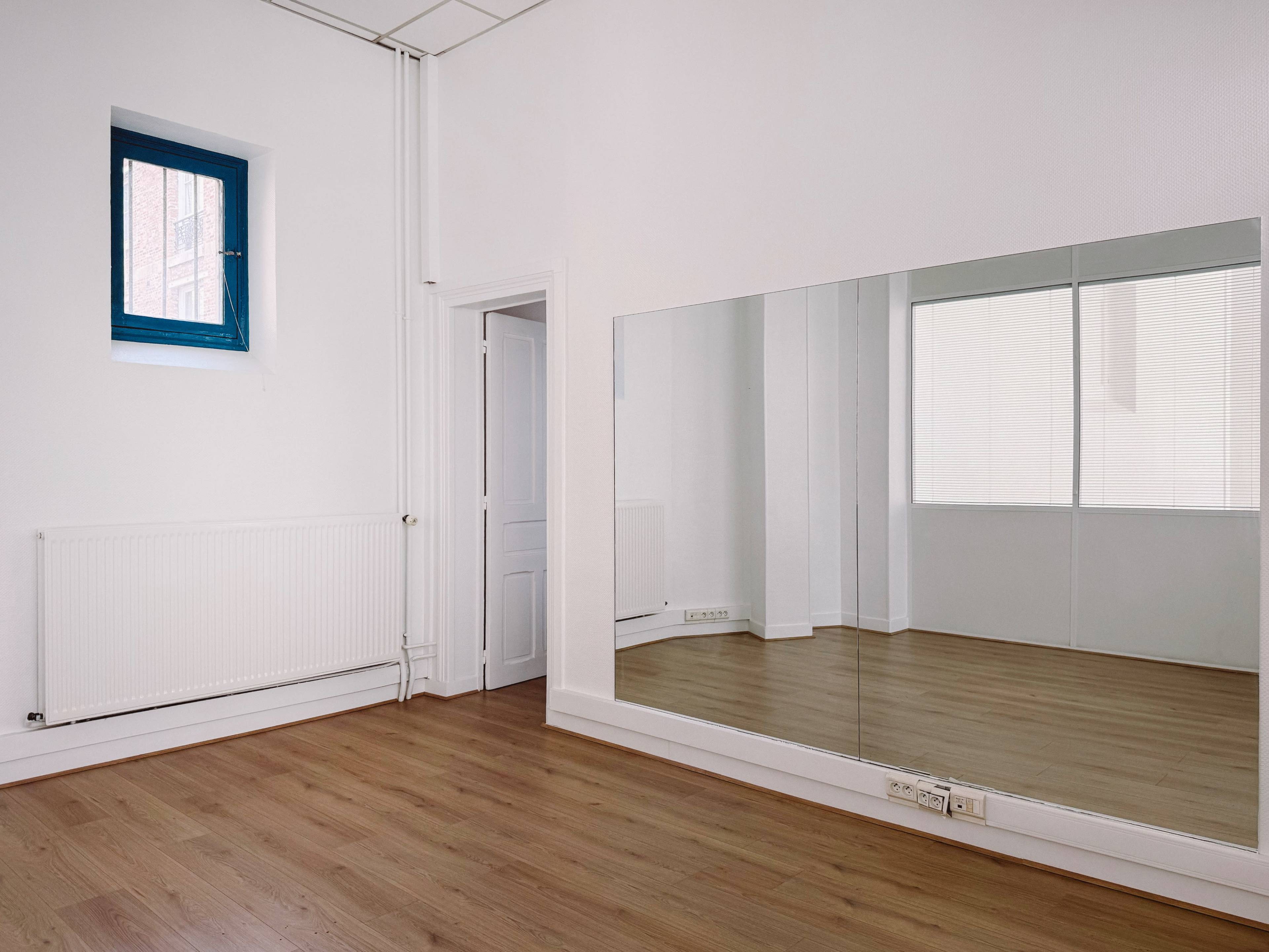 The image shows an empty room with wooden flooring, a large wall mirror, and a window with blue framing.