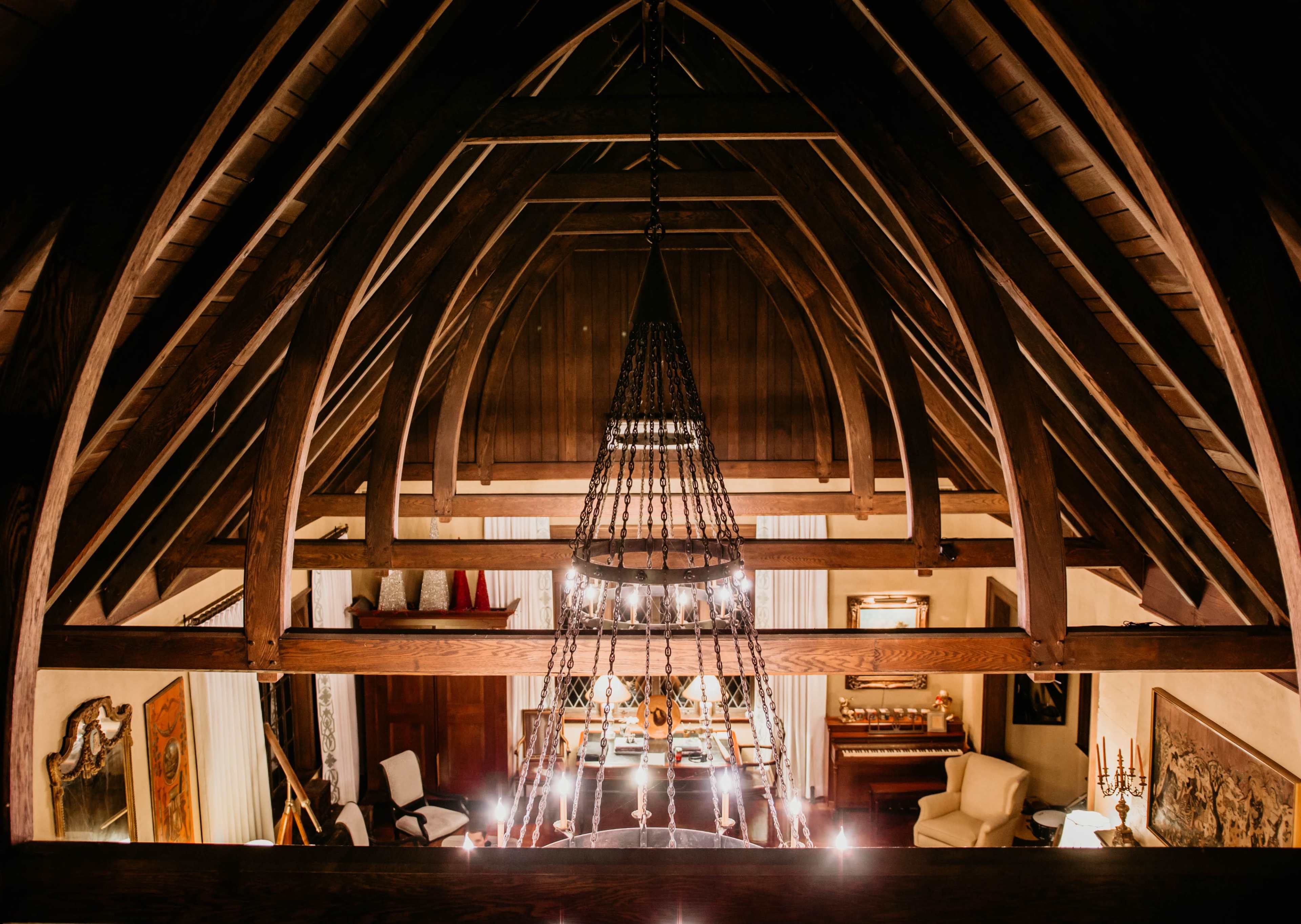 A high ceiling with wooden beams and a chandelier in a spacious room below, featuring various pieces of furniture and decor.