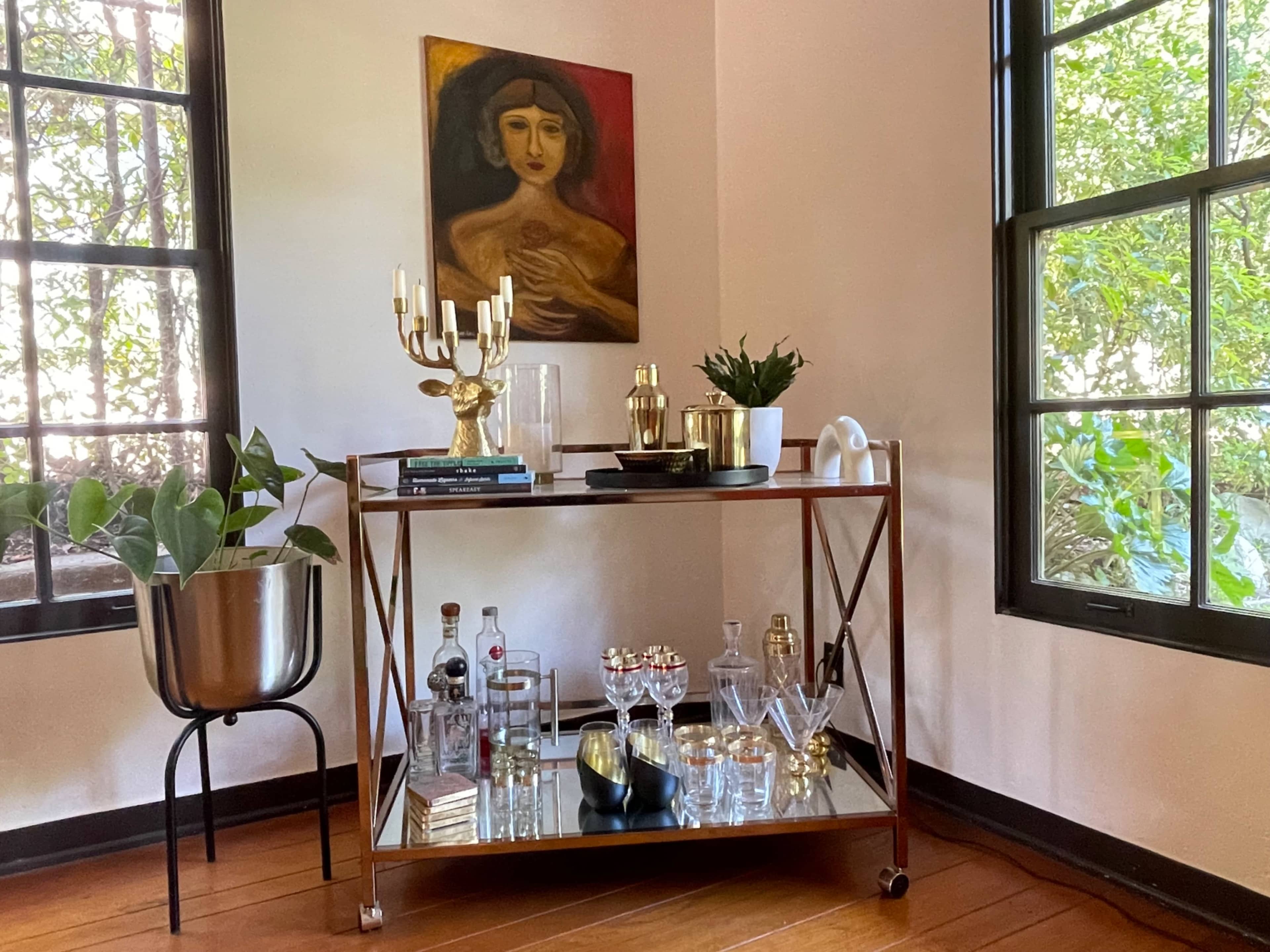 A stylish bar cart is positioned between two windows, featuring various bottles, glassware, and decorative items, with a potted plant and a painting on the wall above.