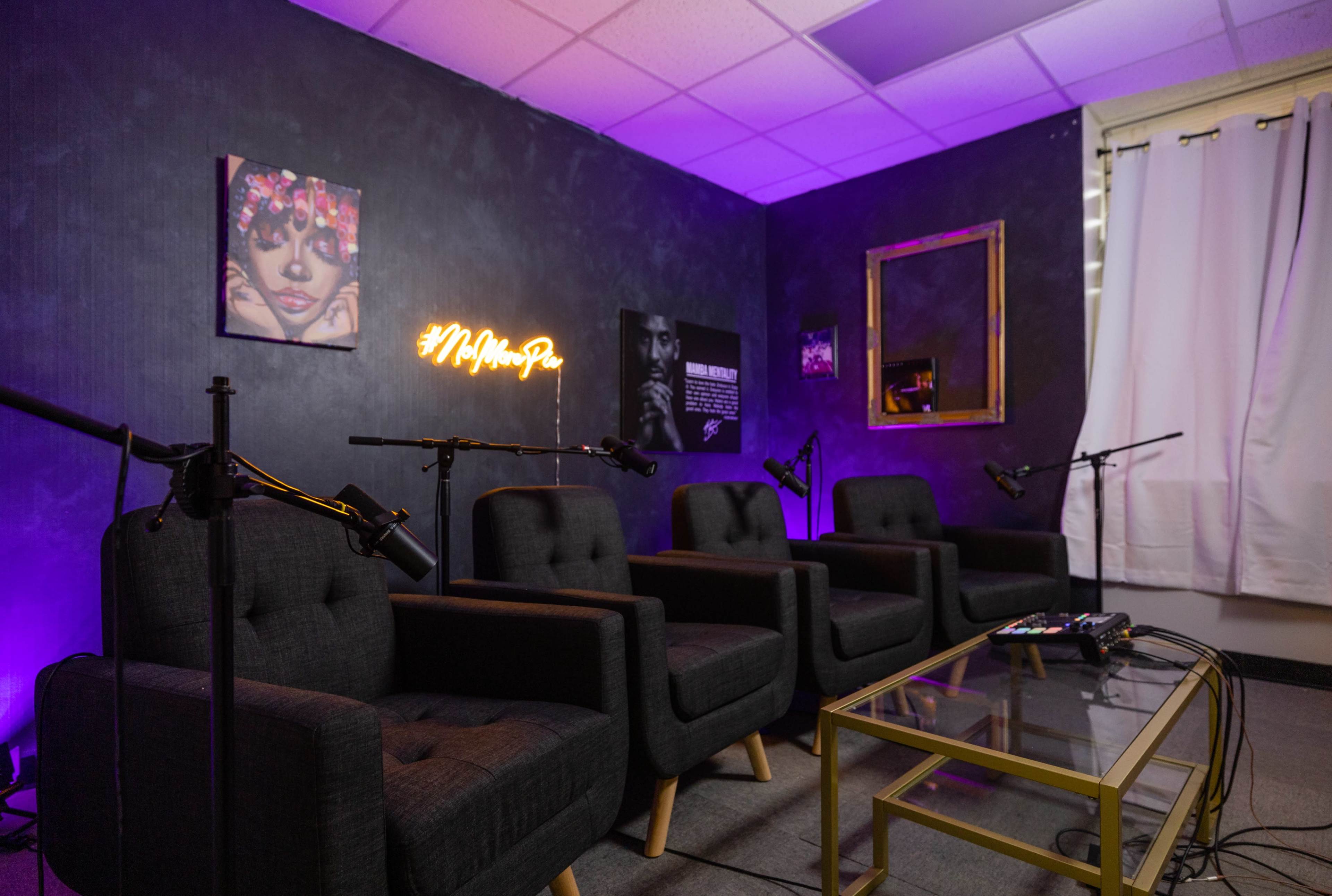 A modern recording studio setup with four dark, upholstered chairs, a glass coffee table, and microphones positioned on stands against a dimly lit wall featuring framed artwork and a illuminated sign.
