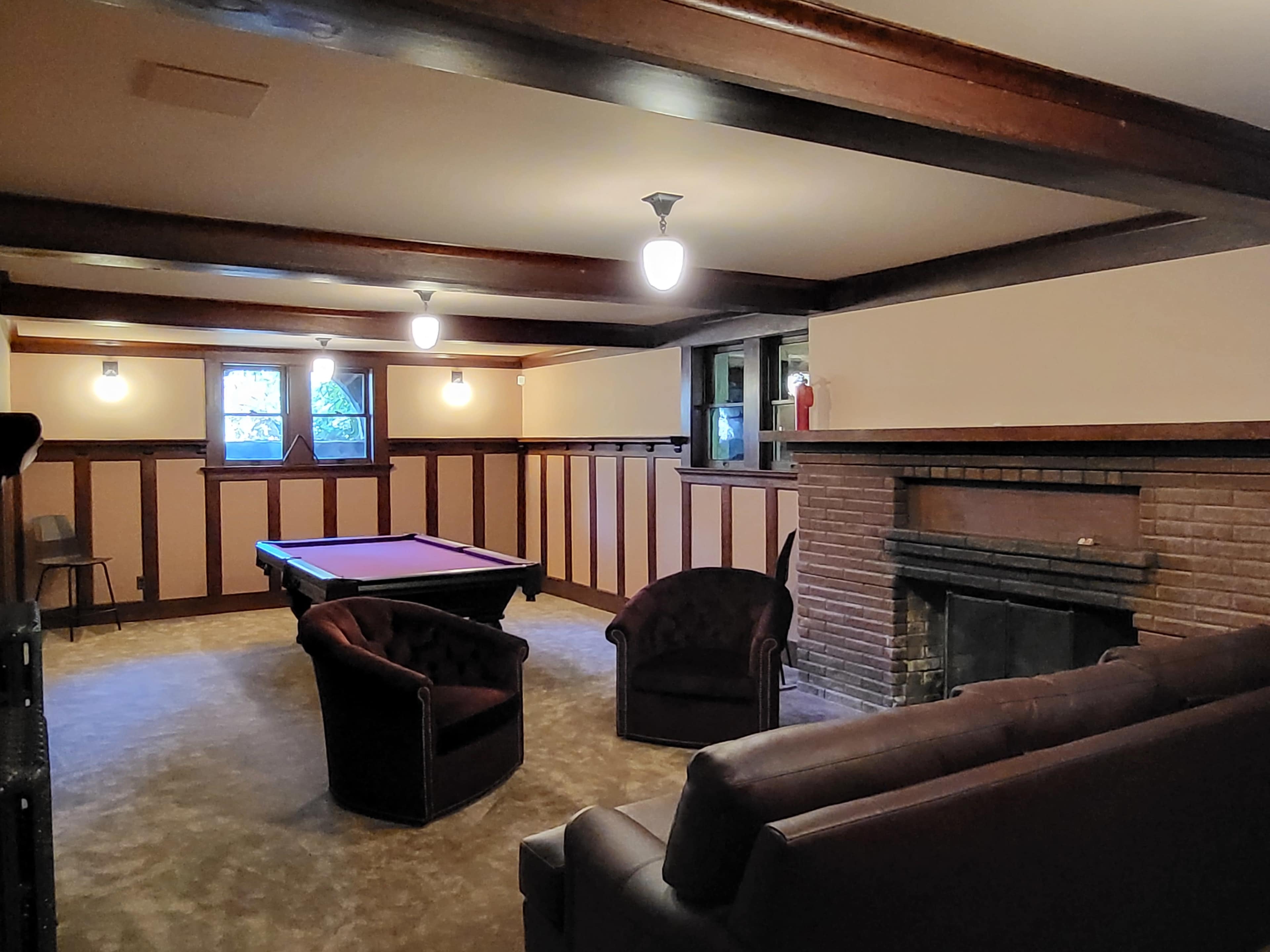 A basement lounge area featuring a pool table, two upholstered chairs, and a fireplace.