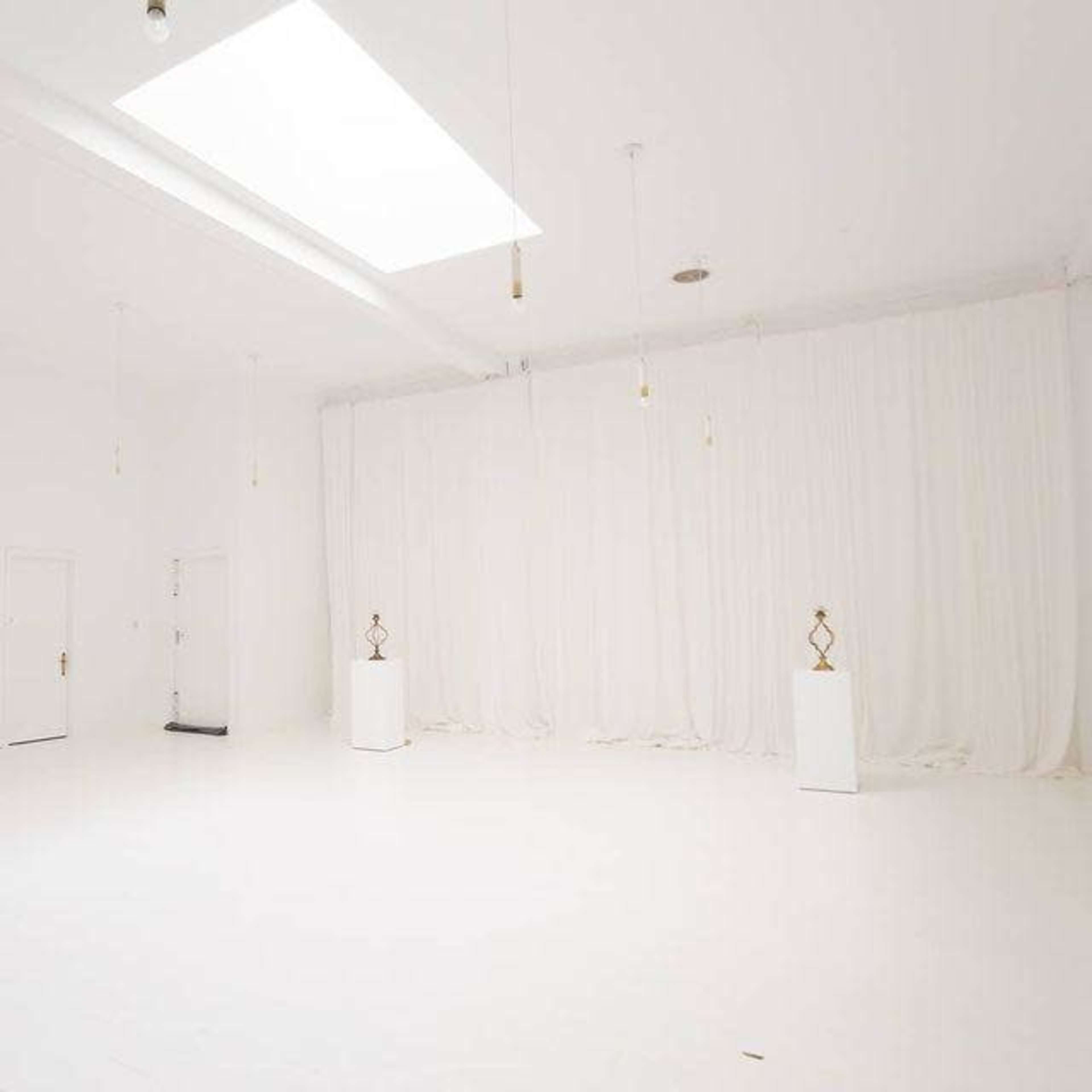 The image shows a spacious, white-walled room with soft white curtains and two bronze sculptures displayed on pedestals.
