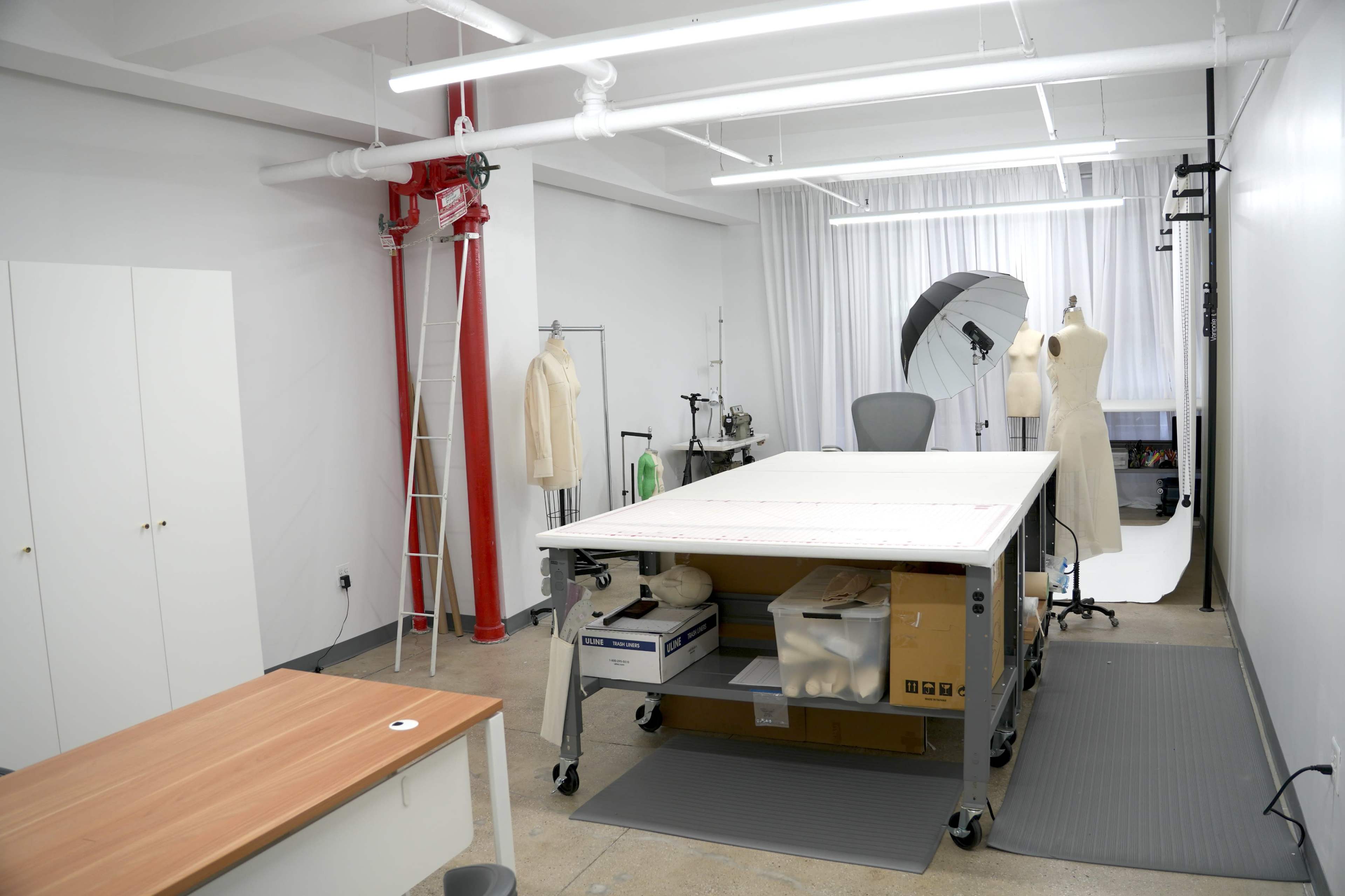 The image shows a spacious design studio with a large table on wheels, a mannequin, and a backdrop setup, along with various tools and materials organized around the room.
