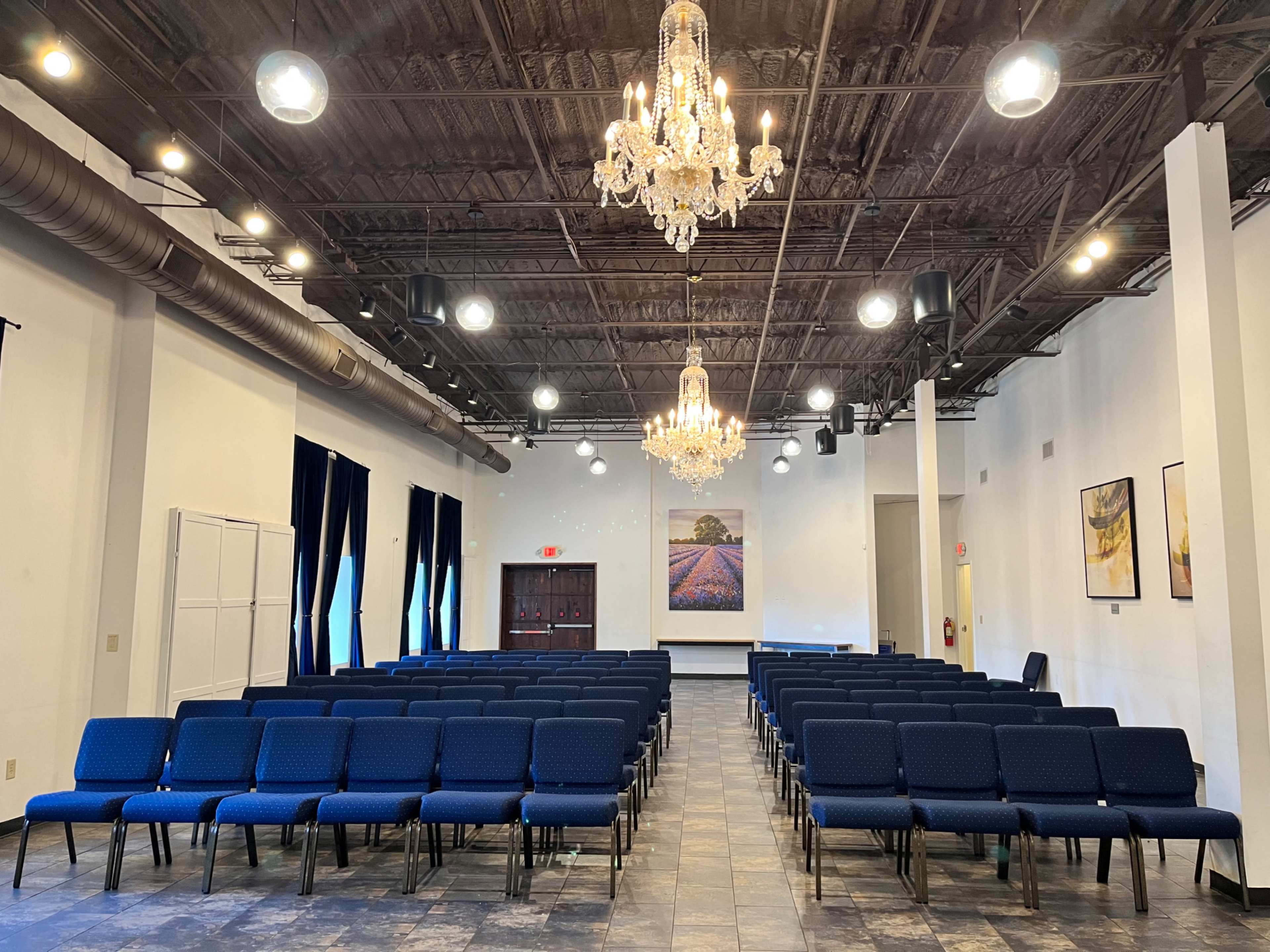 The image shows a spacious room set up with rows of blue chairs facing a stage area illuminated by chandeliers.
