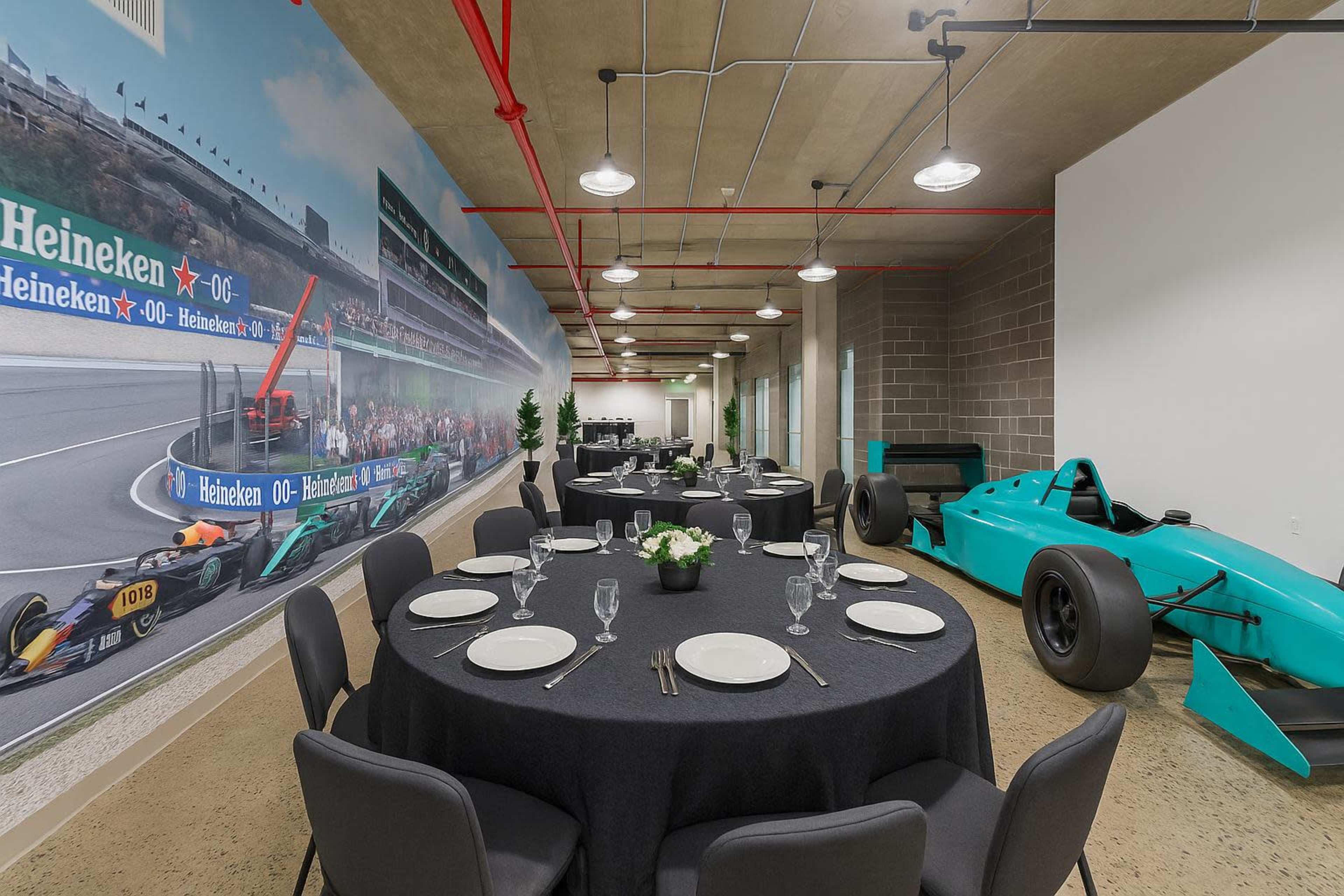 The image shows a modern conference room featuring round tables set for dining and a vintage racing car displayed against a large mural of a racetrack.