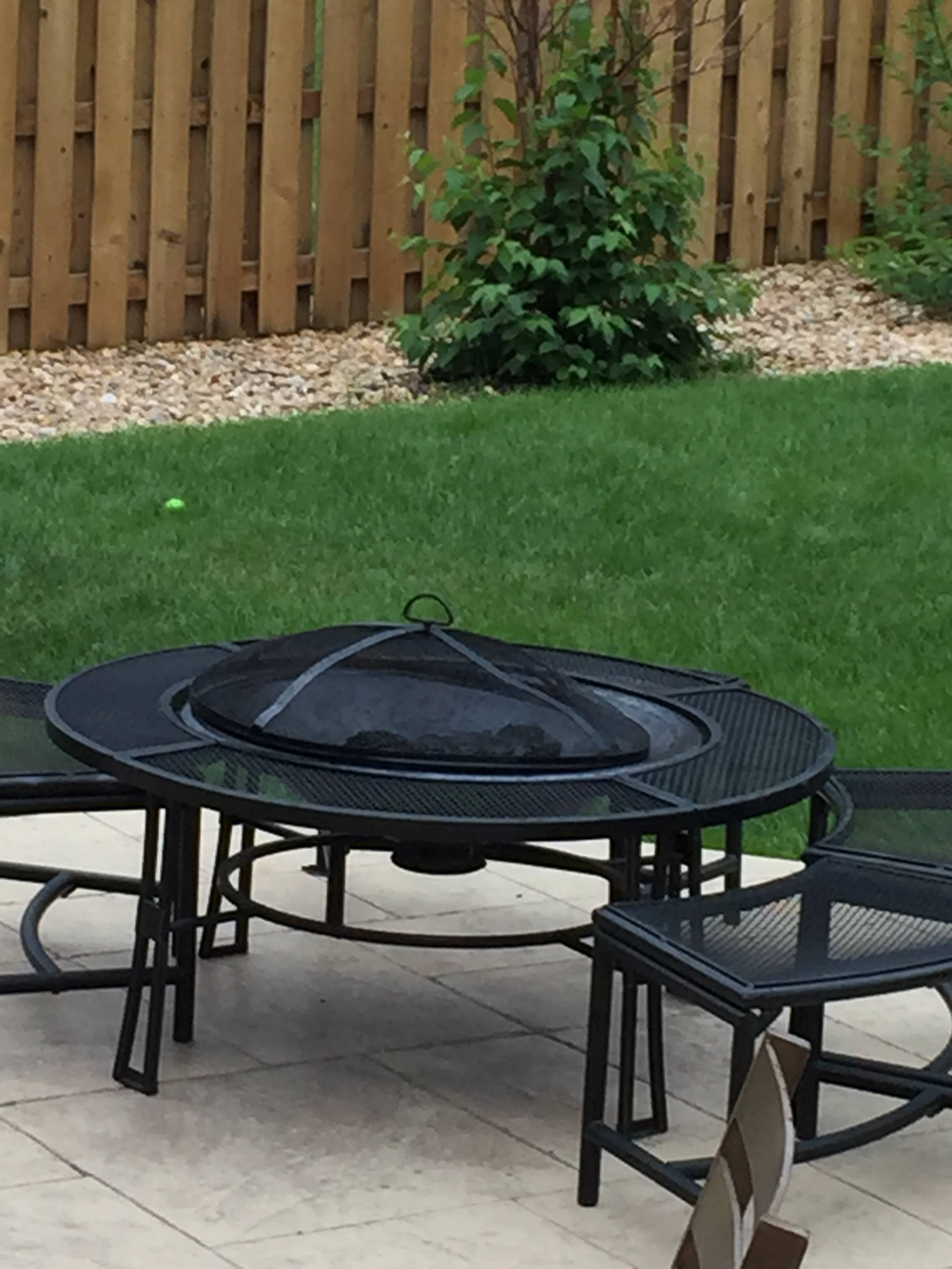A black metal fire pit surrounded by chairs on a stone patio, with grass and a wooden fence in the background.