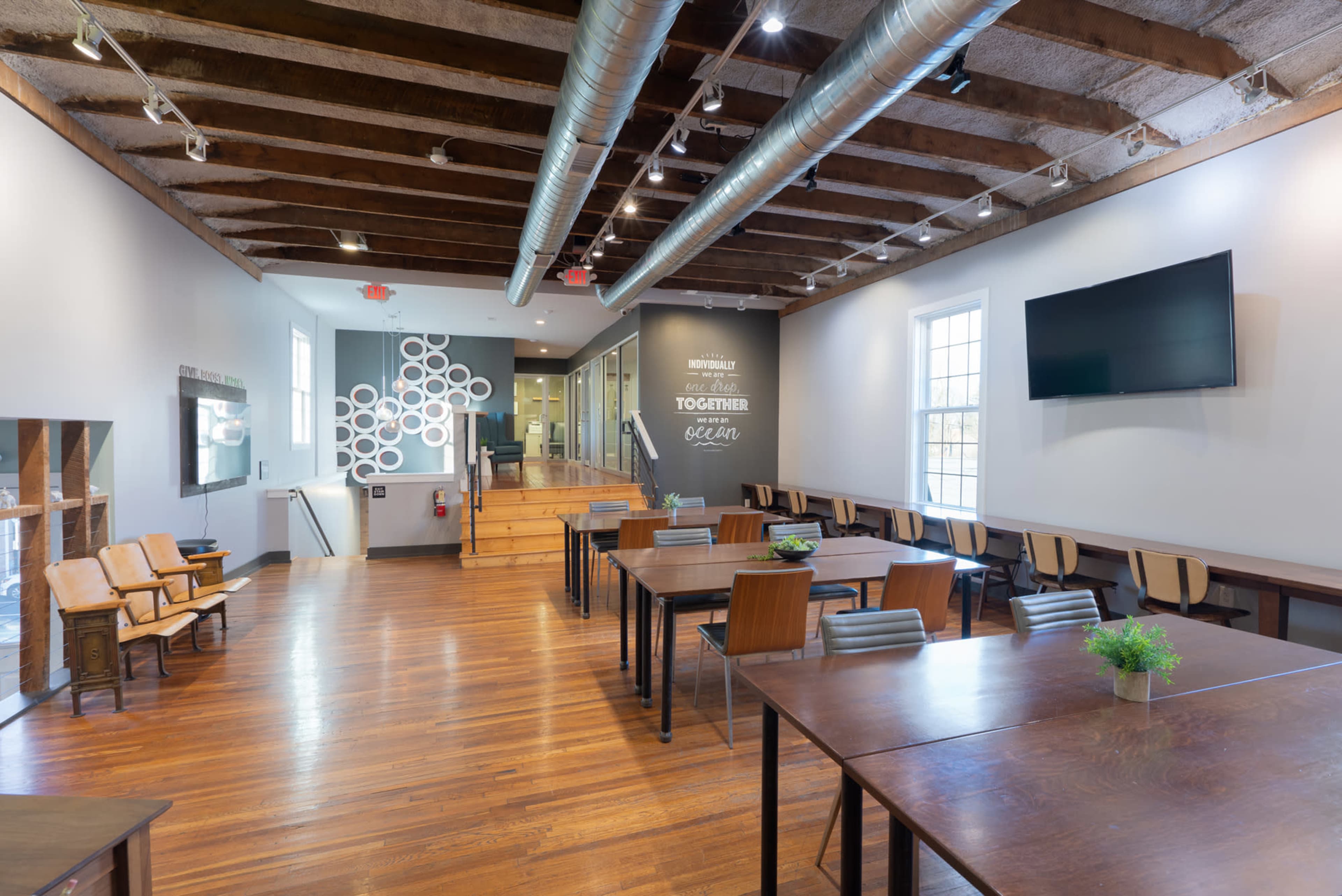 The image shows a spacious meeting room with wooden flooring, large tables set up for seating, and a wall with inspirational text, illuminated by natural light from the windows.