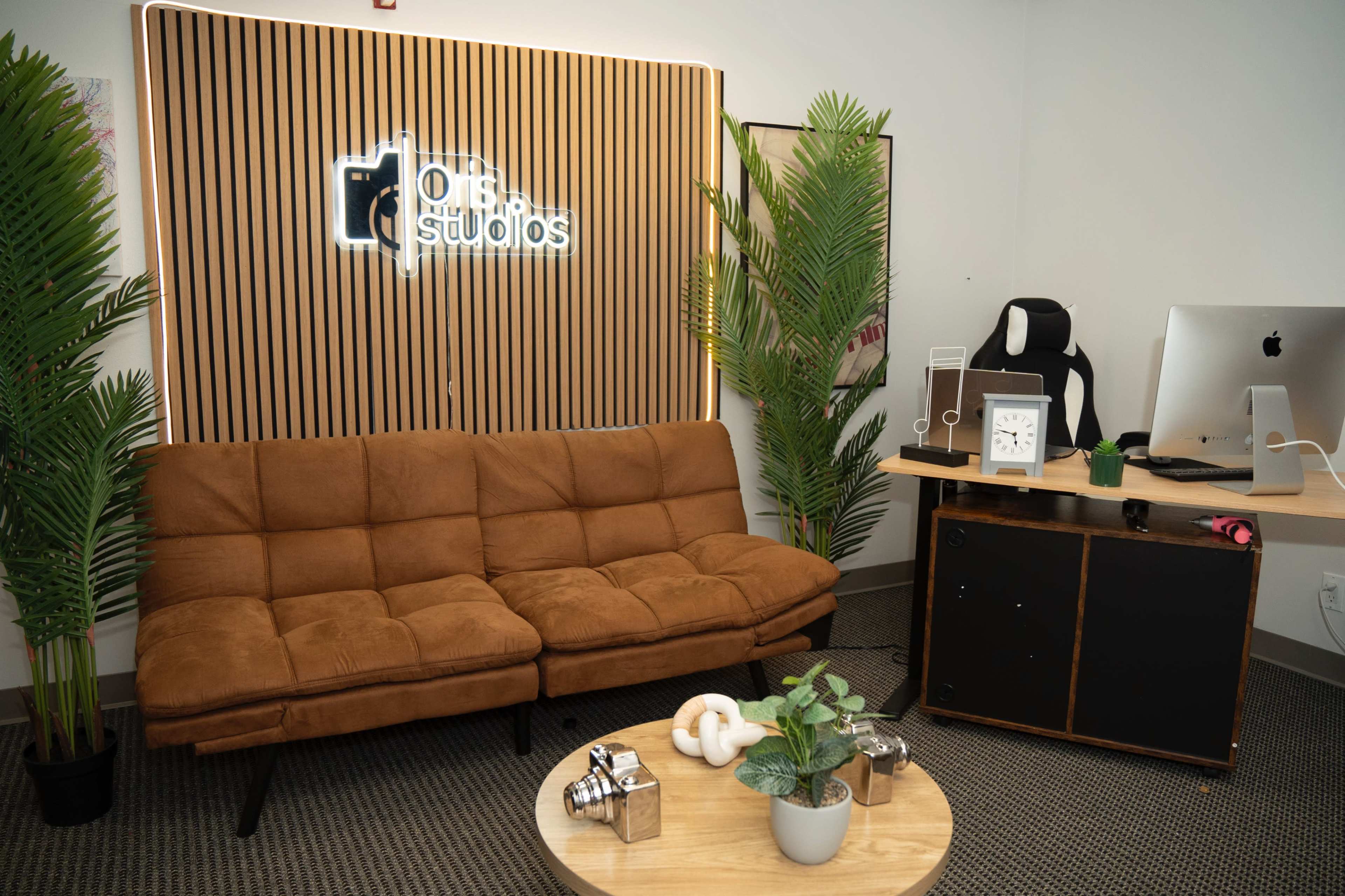 The image shows a modern office space featuring a brown couch, a round coffee table with decorative items, and a desk with a computer and an ergonomic chair, surrounded by indoor plants.