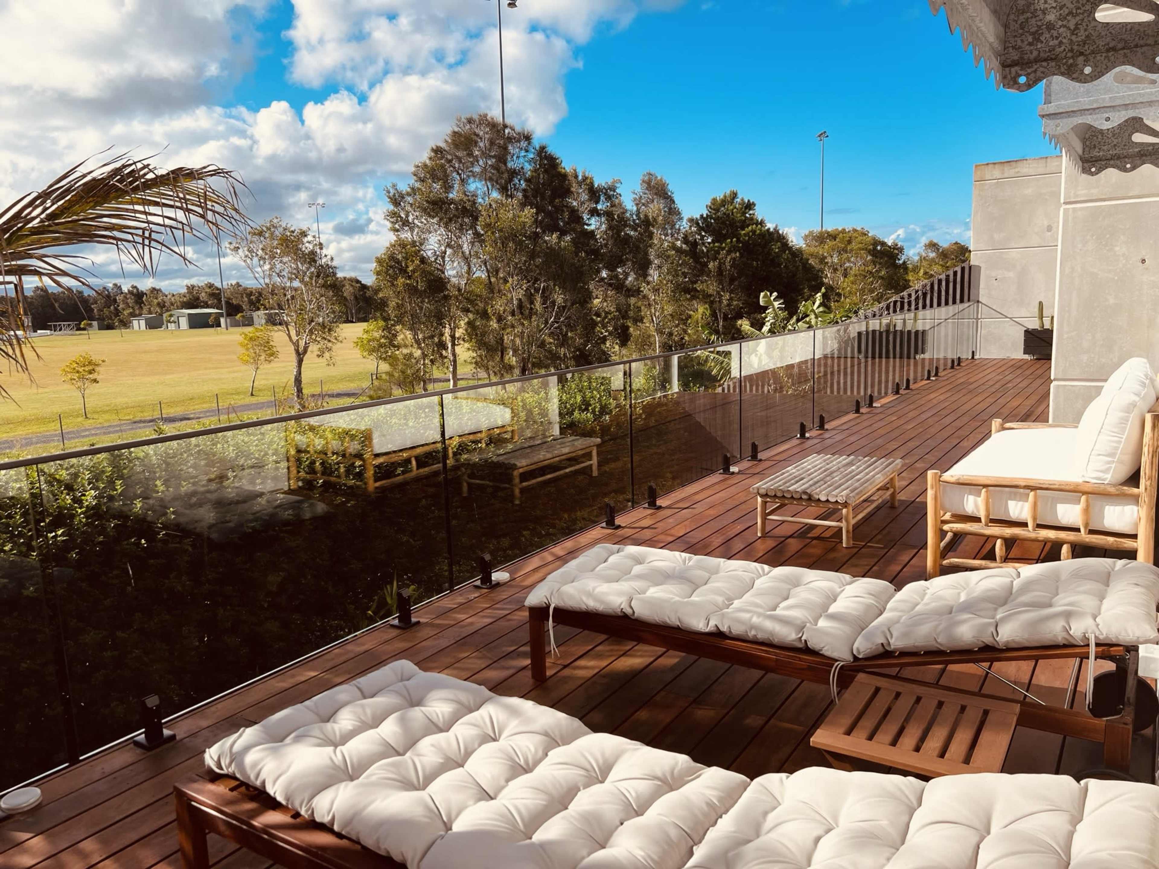 A wooden deck with white lounge chairs and a glass railing overlooks a grassy field and trees under a partly cloudy sky.
