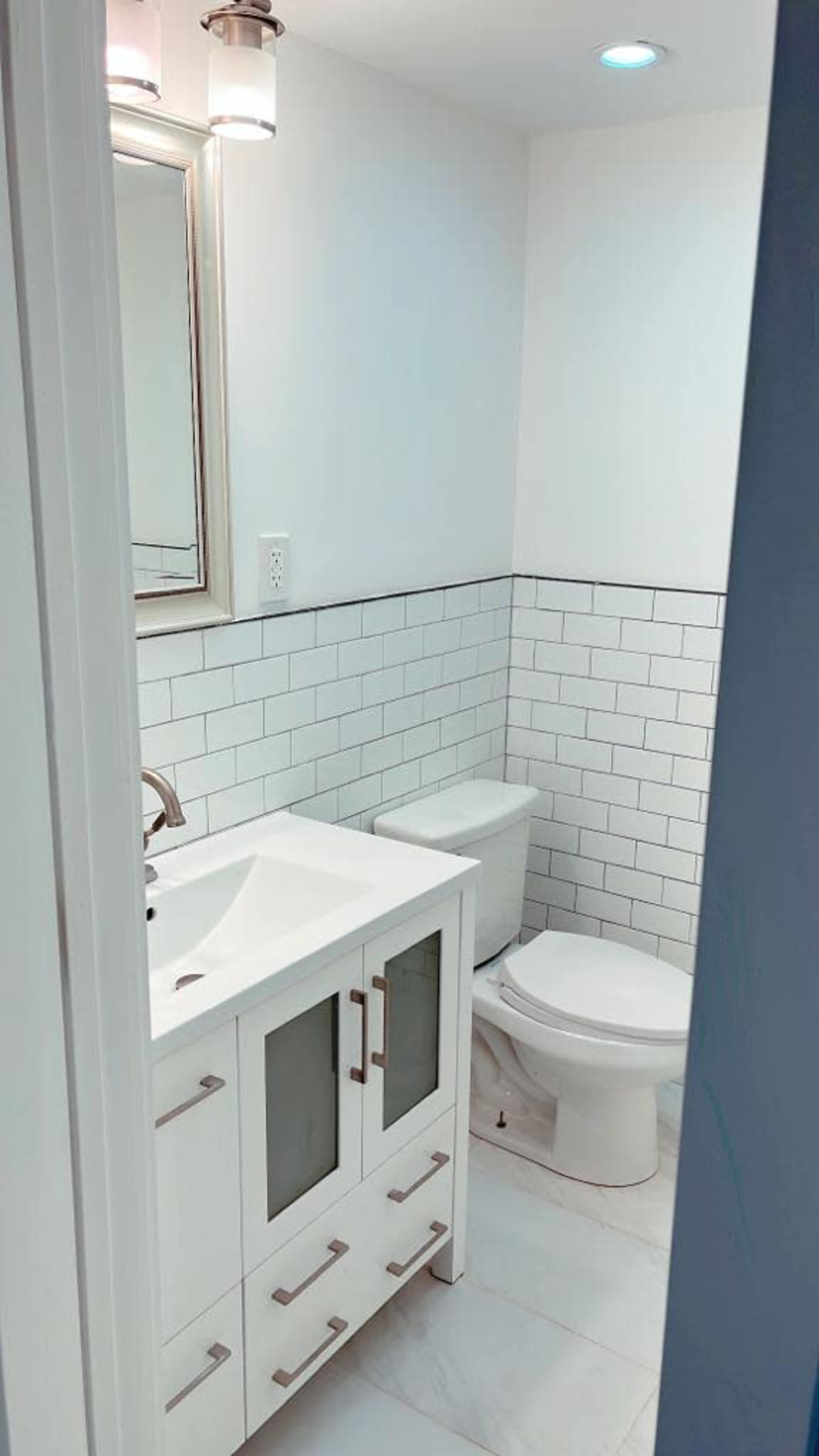 The image shows a small bathroom with a white vanity featuring glass doors, a mirror above it, a toilet, and white tiled walls.