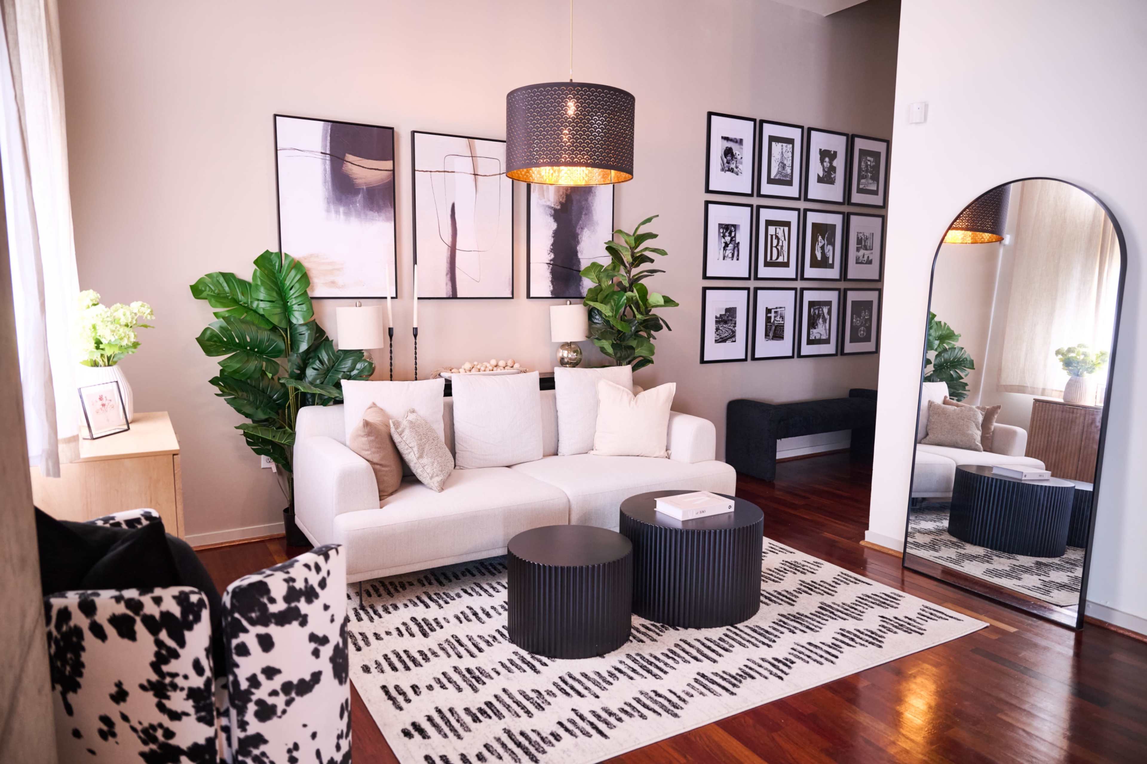 The living room features a light-colored sofa with decorative pillows, two round black coffee tables, framed black-and-white photos on the wall, and large green plants.