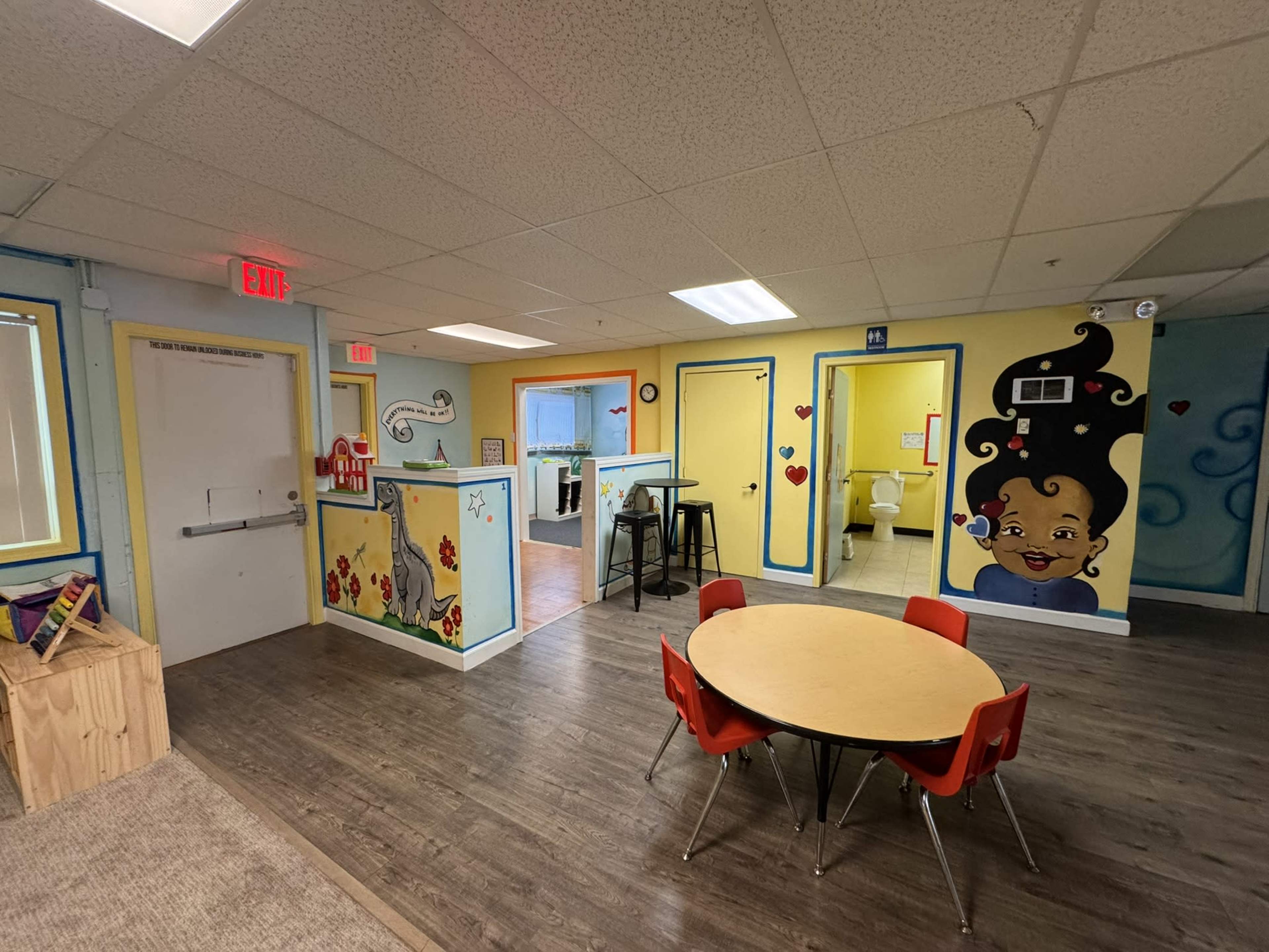 The image shows a brightly colored classroom with murals on the walls, a round table with red chairs, and a view of adjacent rooms including a rest area.