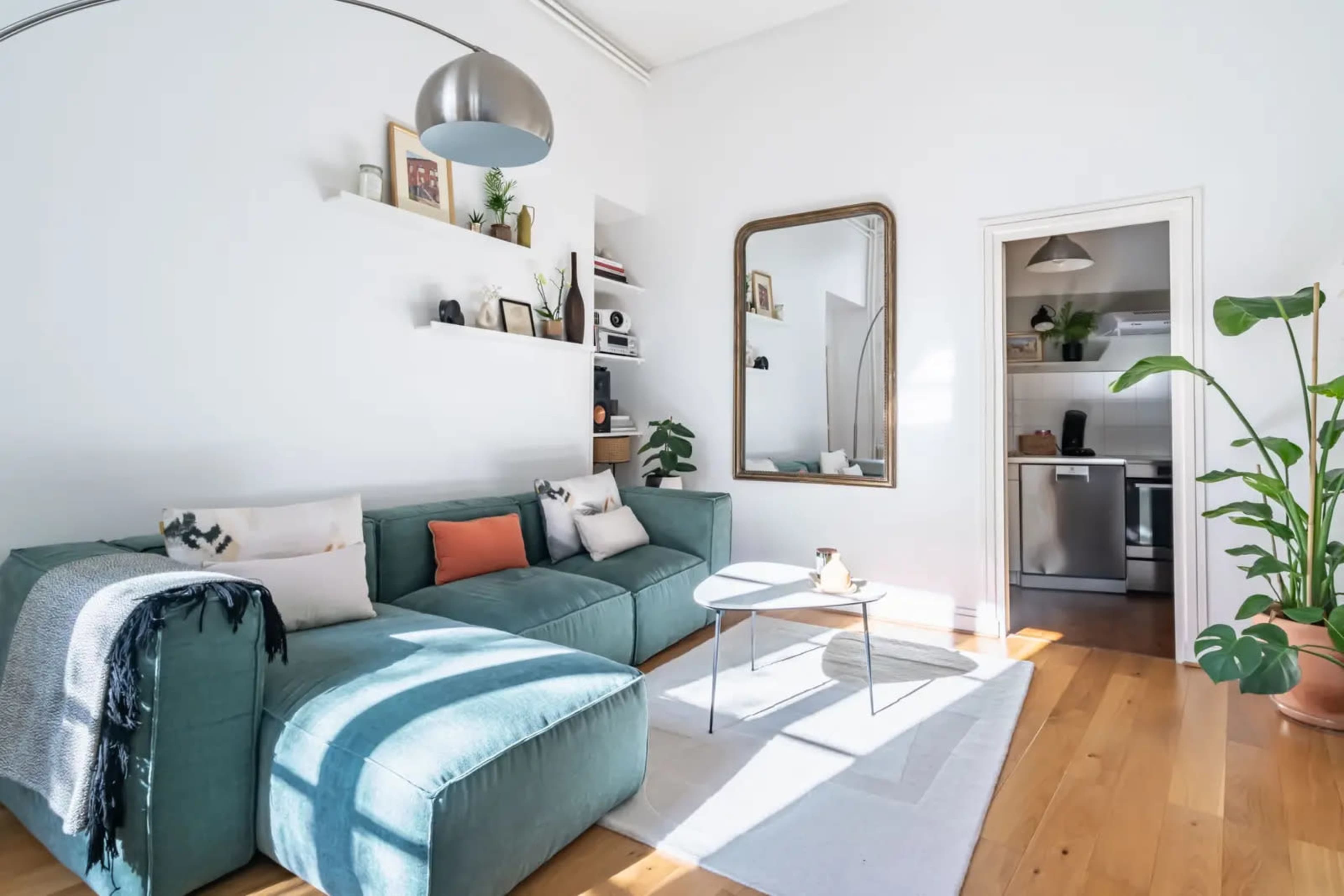 The image shows a modern living room featuring a teal sectional sofa, a round coffee table, and a large mirror on the wall opposite a kitchen area.