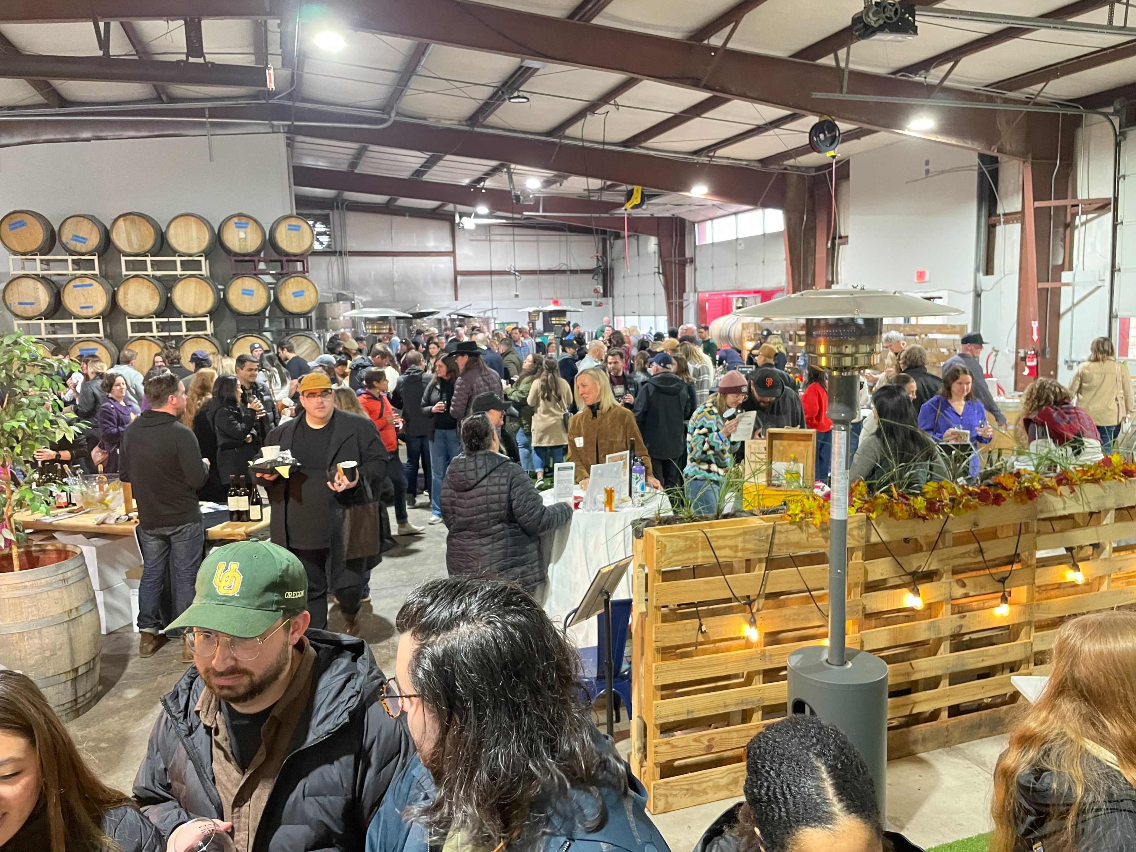 A large crowd gathers inside a warehouse-like space filled with food vendors, wooden barrels, and seasonal decorations.