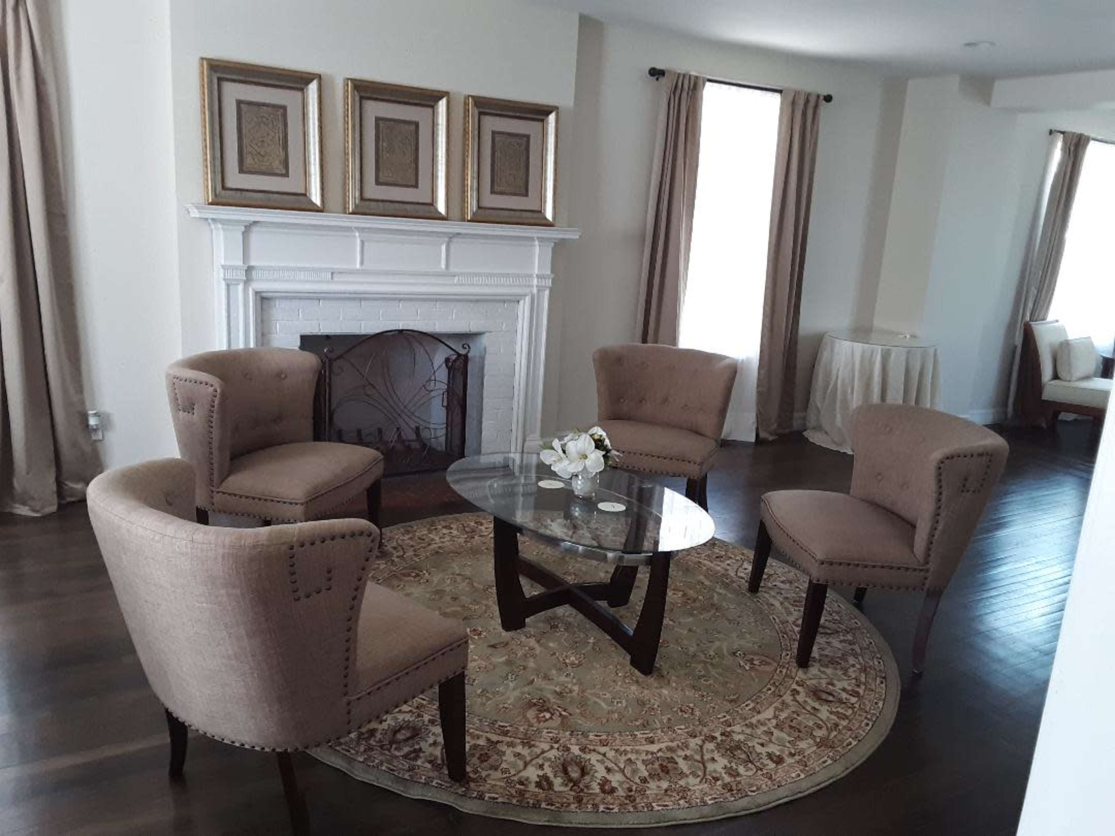 A modern living room with four beige upholstered chairs arranged around a glass coffee table on a patterned rug, in front of a fireplace decorated with three framed artworks.