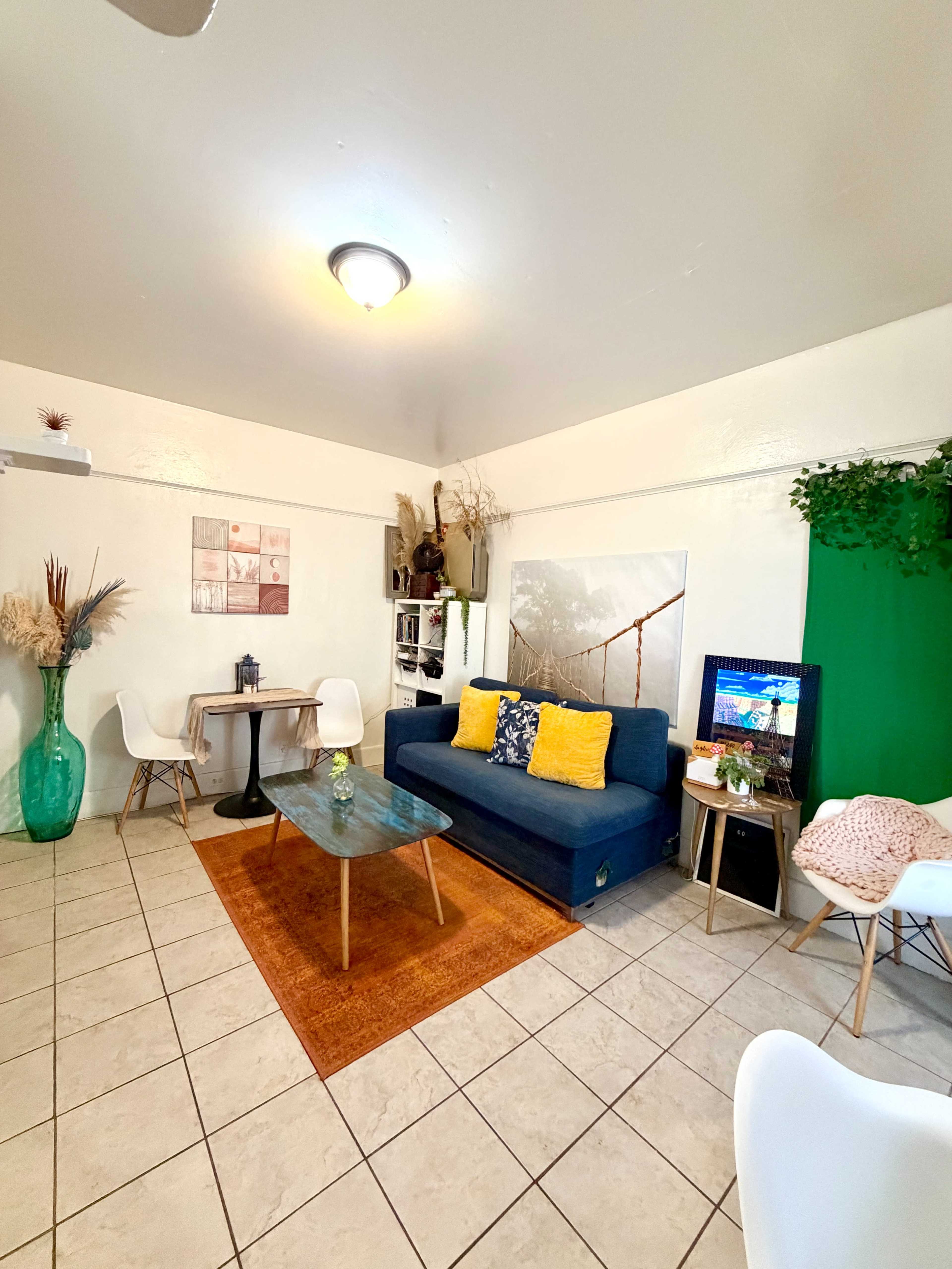 The image shows a cozy living room with a blue sofa, an orange rug, a small dining table, and decorative plants.