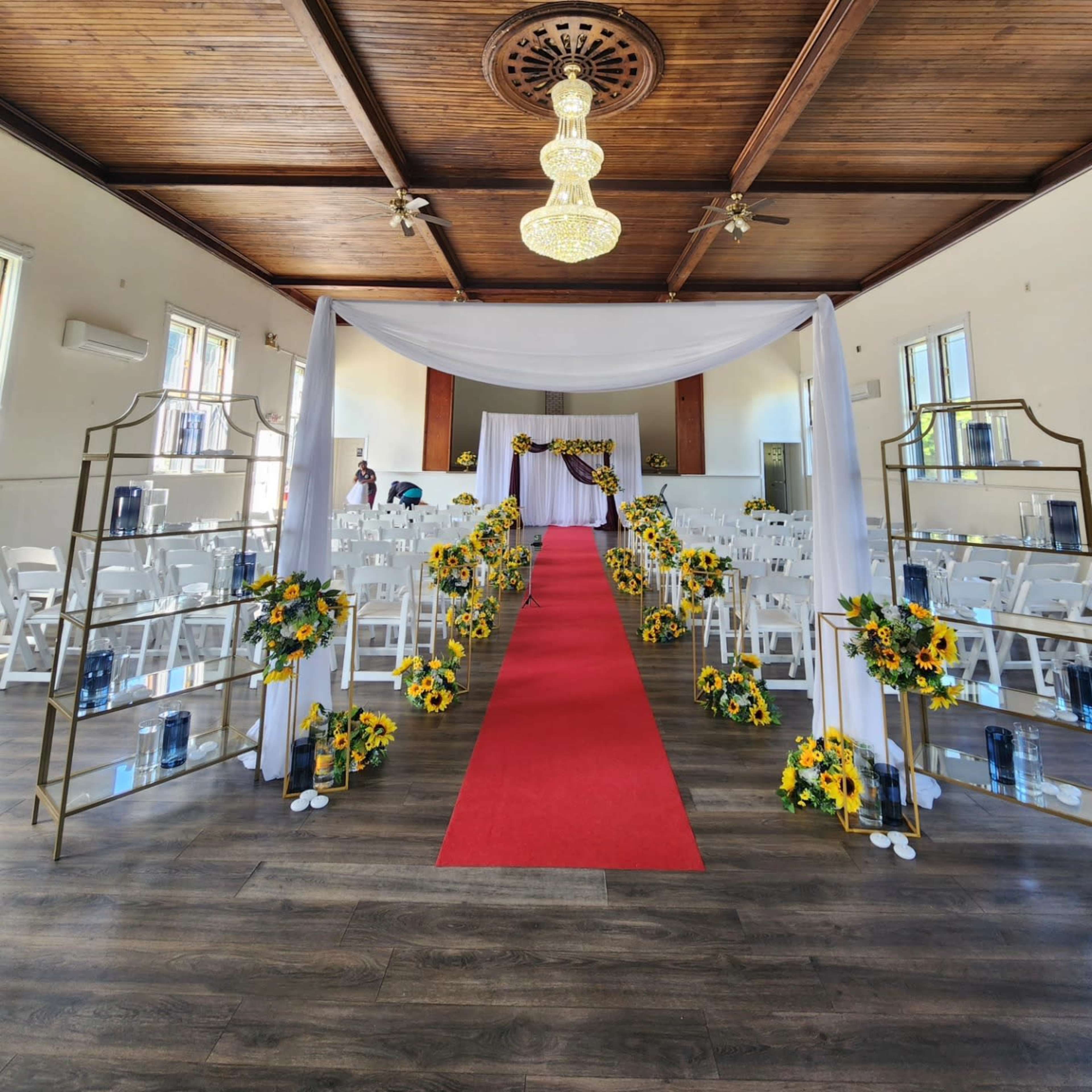 The image shows a decorated indoor space set up for a wedding ceremony, featuring a red carpet, floral arrangements, and chairs neatly placed on either side.