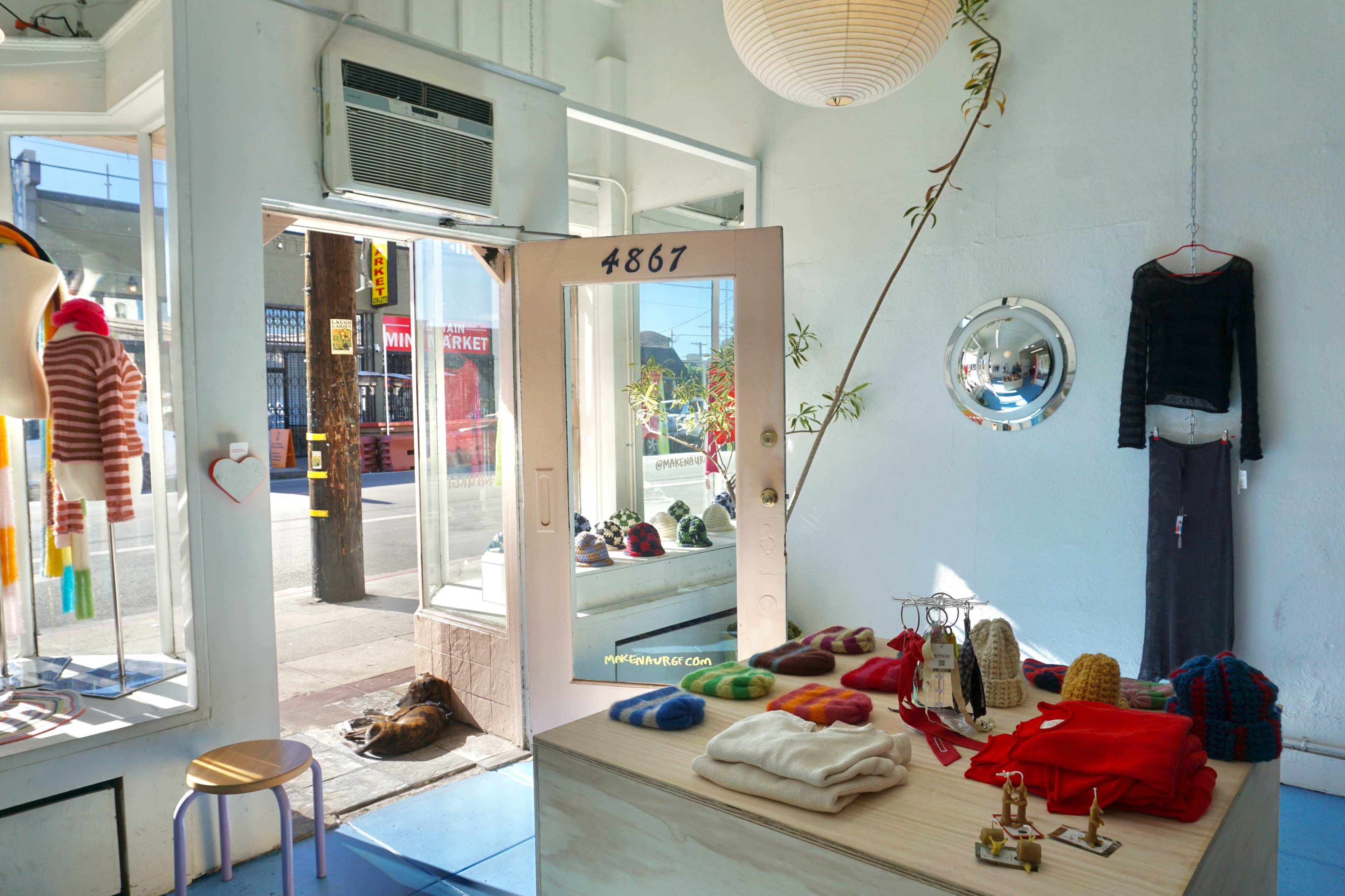 The image shows a boutique interior with colorful clothing items displayed on a wooden table, large windows letting in sunlight, and an open door leading to the street.