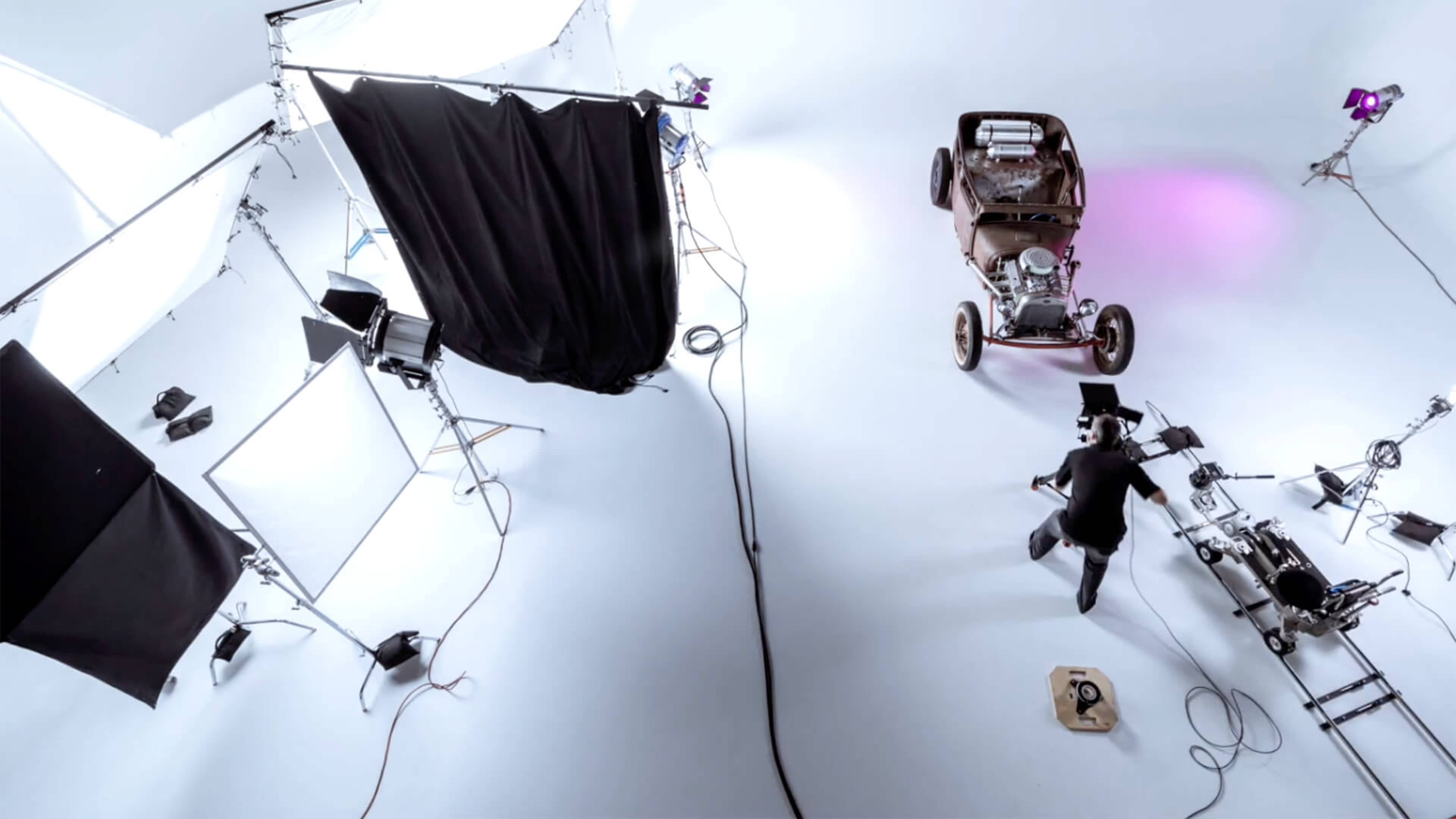 A studio set is arranged with various lighting setups, a black curtain, and a vintage car positioned on a light-colored floor.