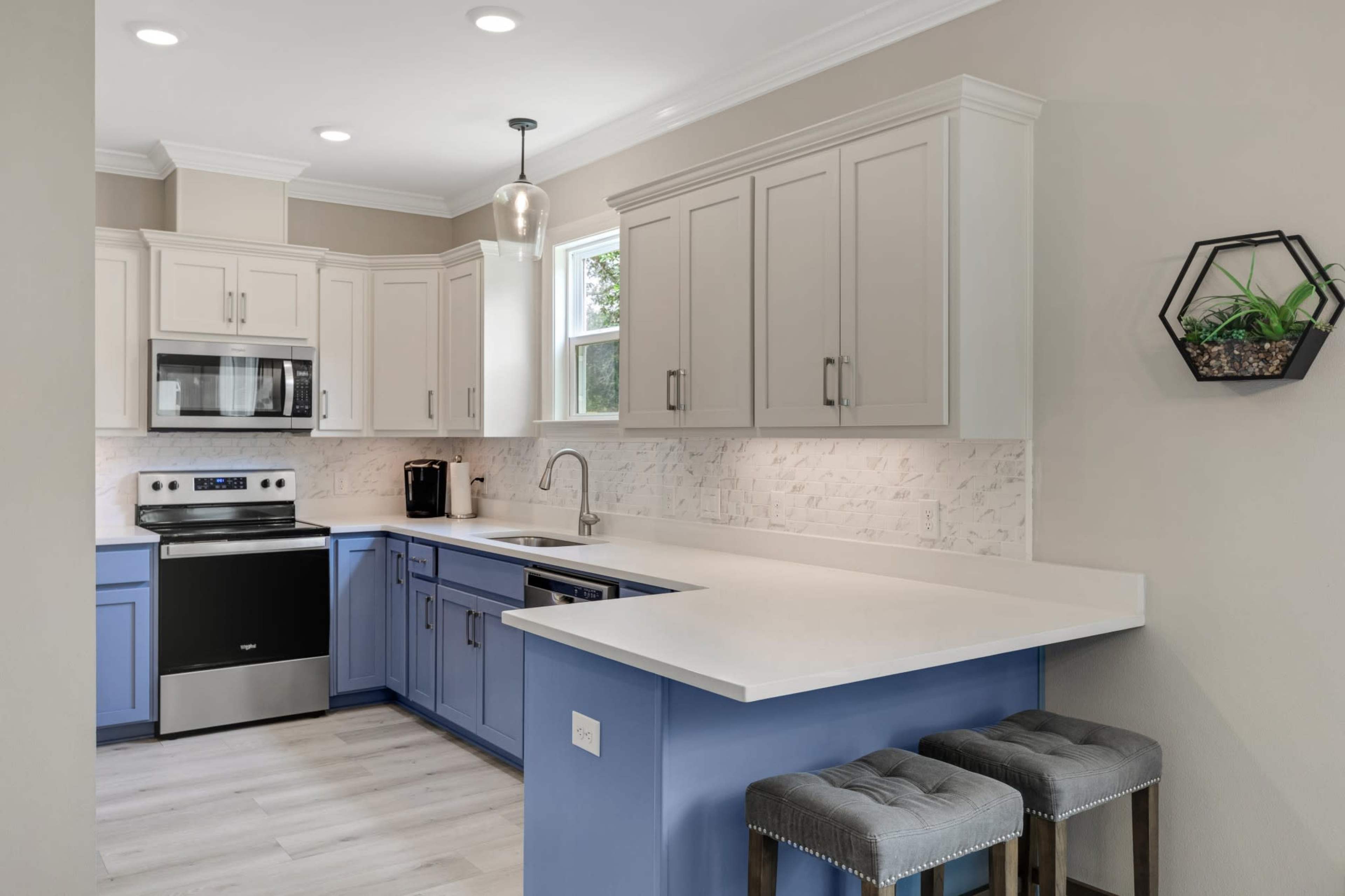 The kitchen features a combination of light gray and blue cabinetry, stainless steel appliances, and a white countertop with two gray stools at the bar.