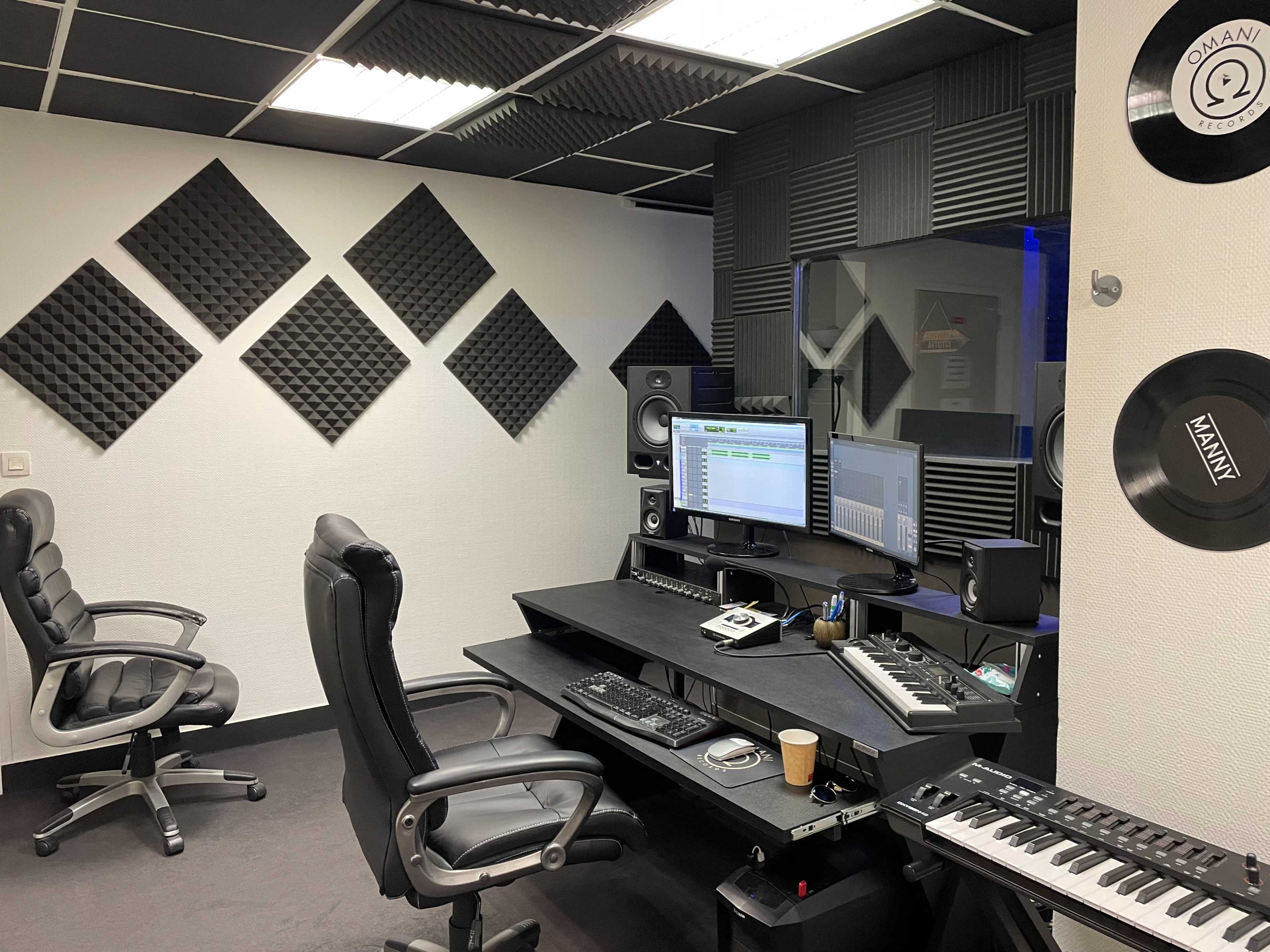 A soundproofed music studio featuring a black desk with computer monitors, audio equipment, and two ergonomic chairs.