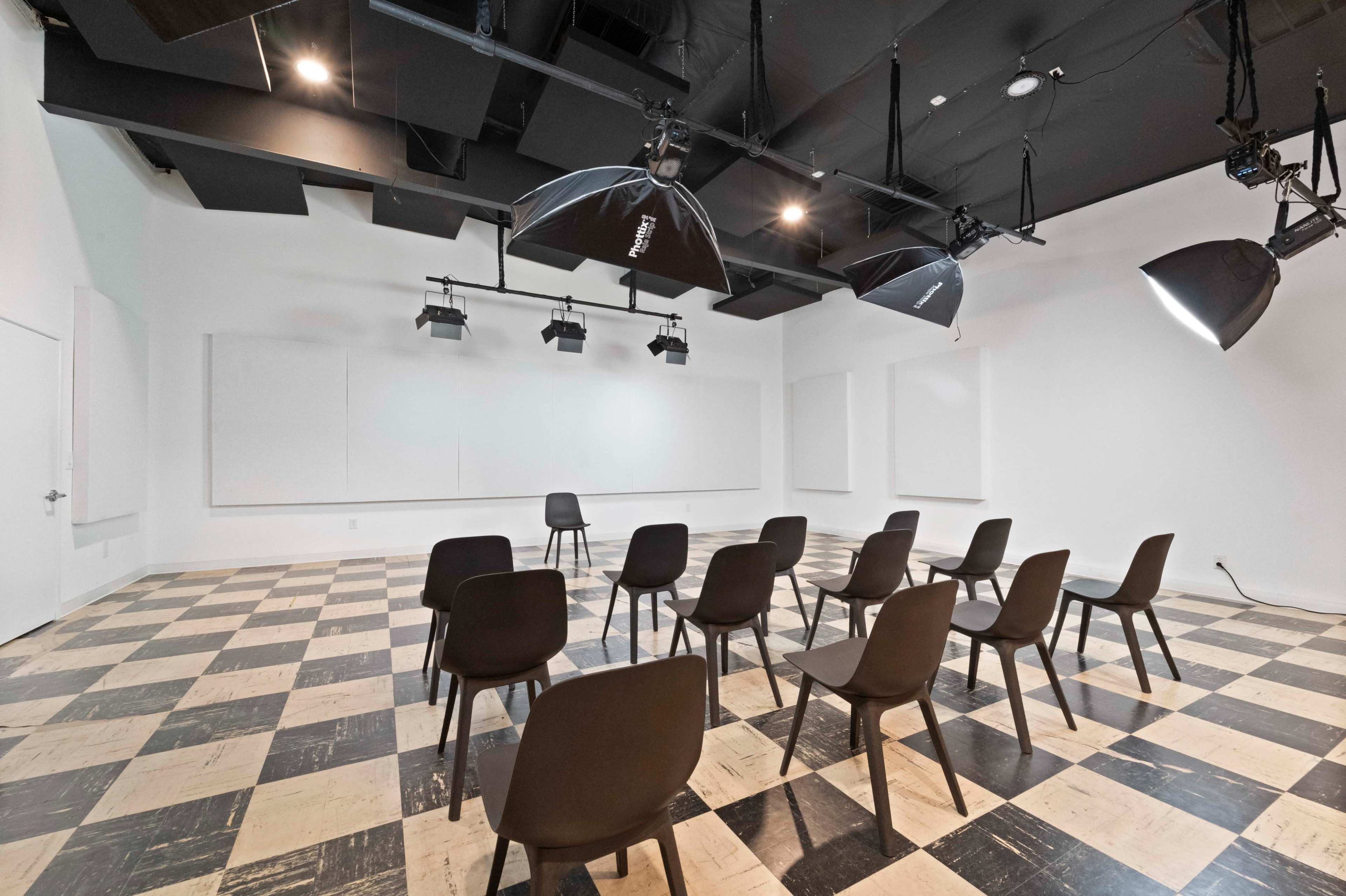 A minimalistic conference room with checkered flooring, black chairs arranged in rows, and overhead lighting fixtures.