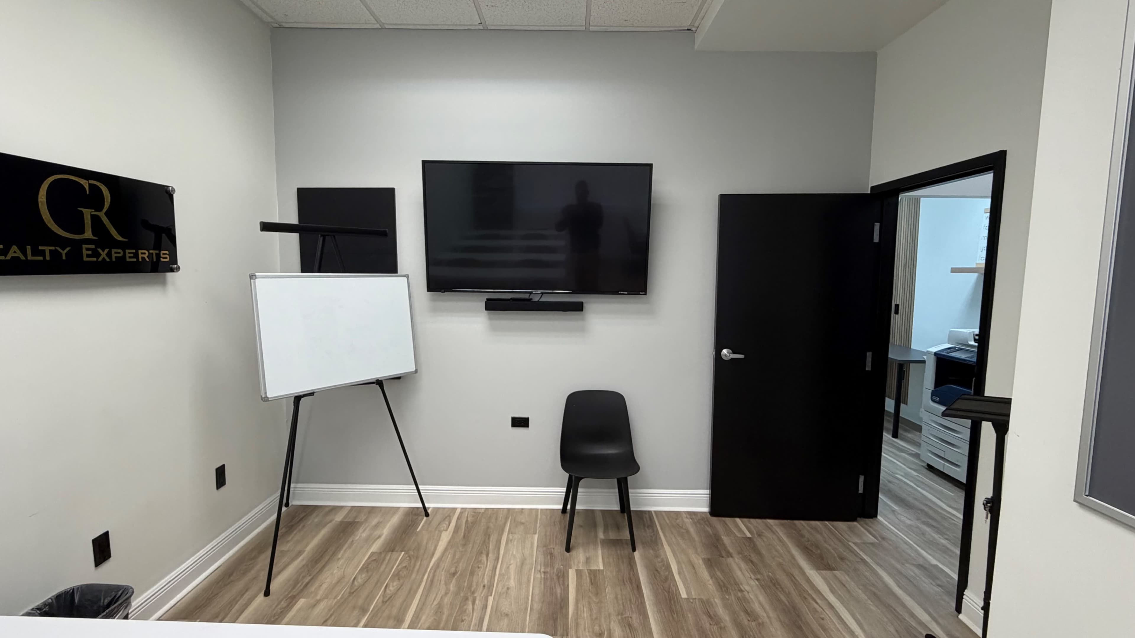 The image shows a small, modern meeting room with a wall-mounted television, a black chair, an easel with a whiteboard, and an open door leading to another room.