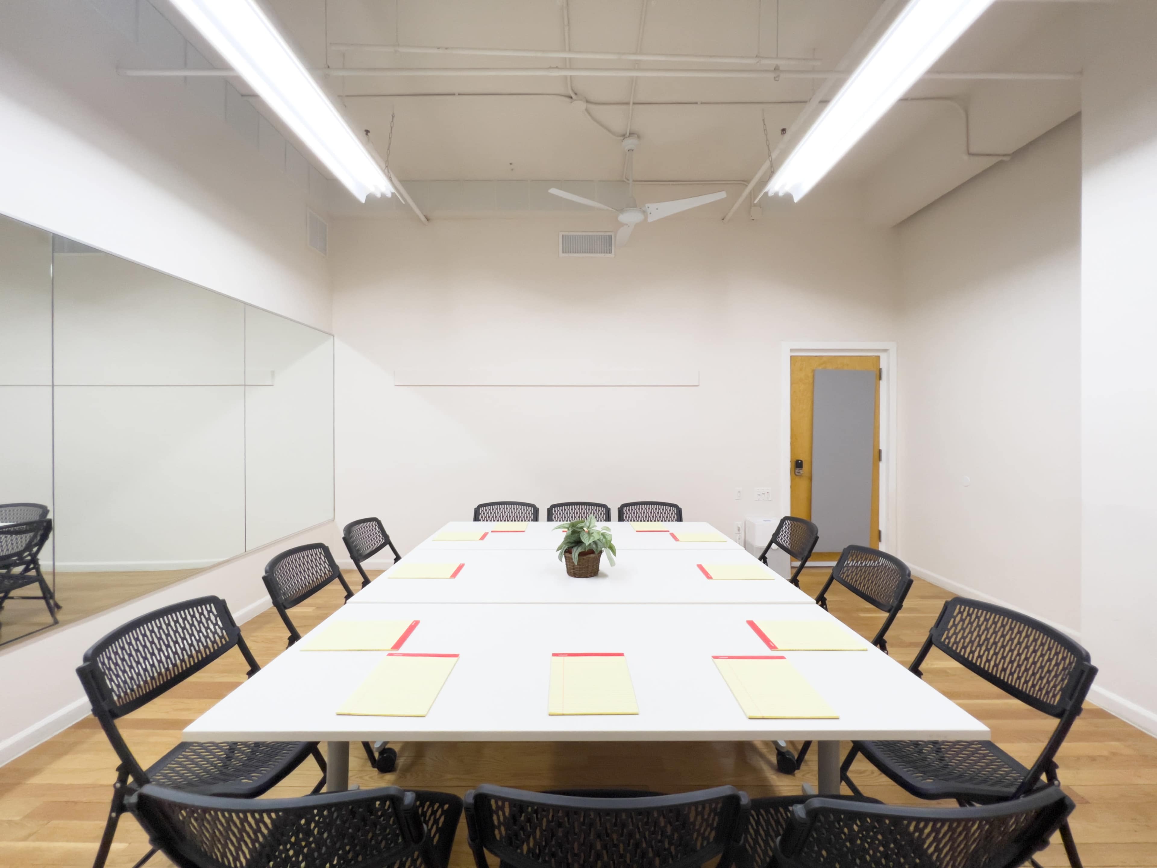 A conference room features a long table surrounded by black chairs, with yellow papers and a small plant at the center.