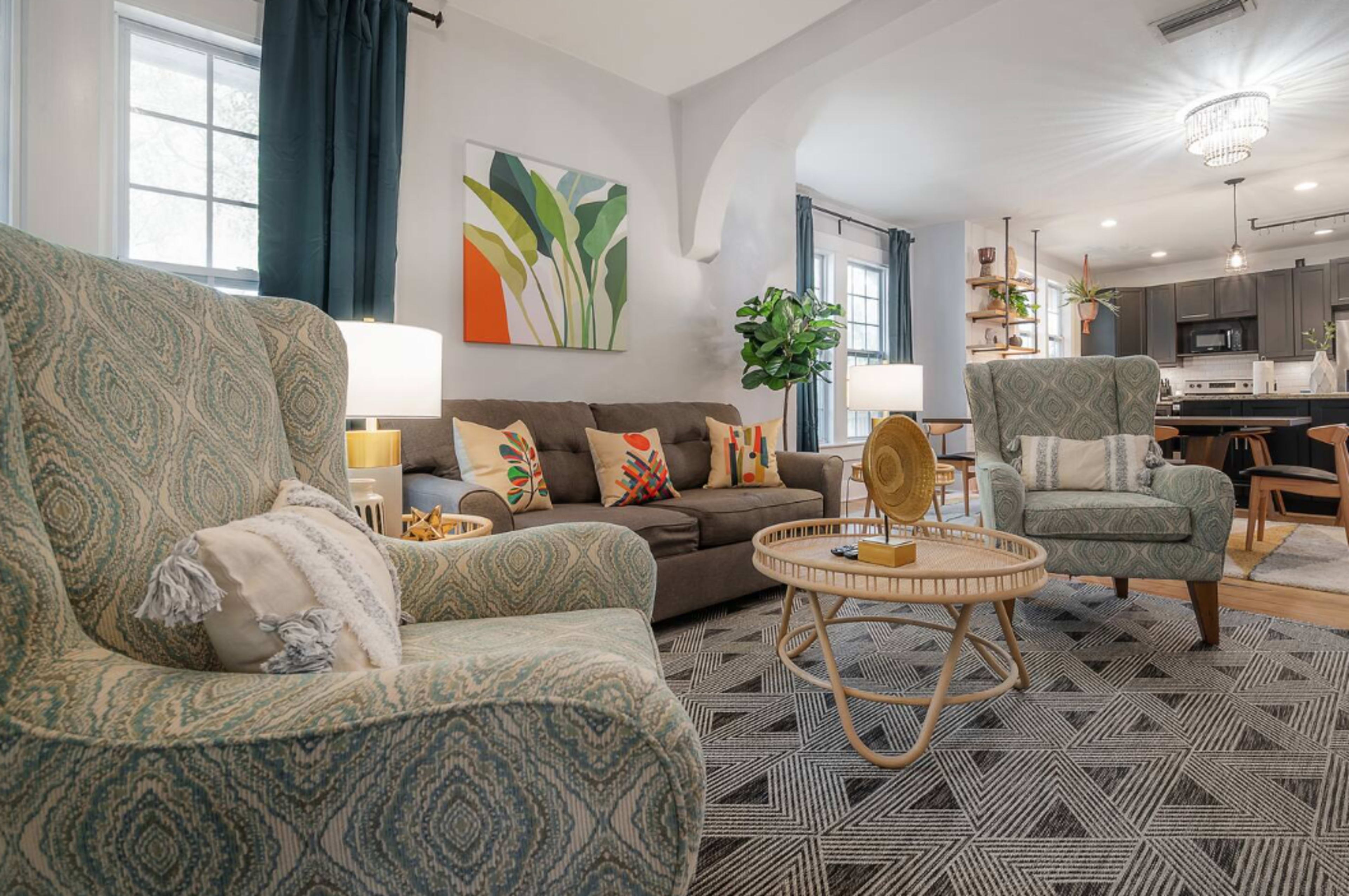 A cozy living room featuring two upholstered chairs, a sofa with decorative pillows, a circular coffee table, and a large plant, with the kitchen in the background.