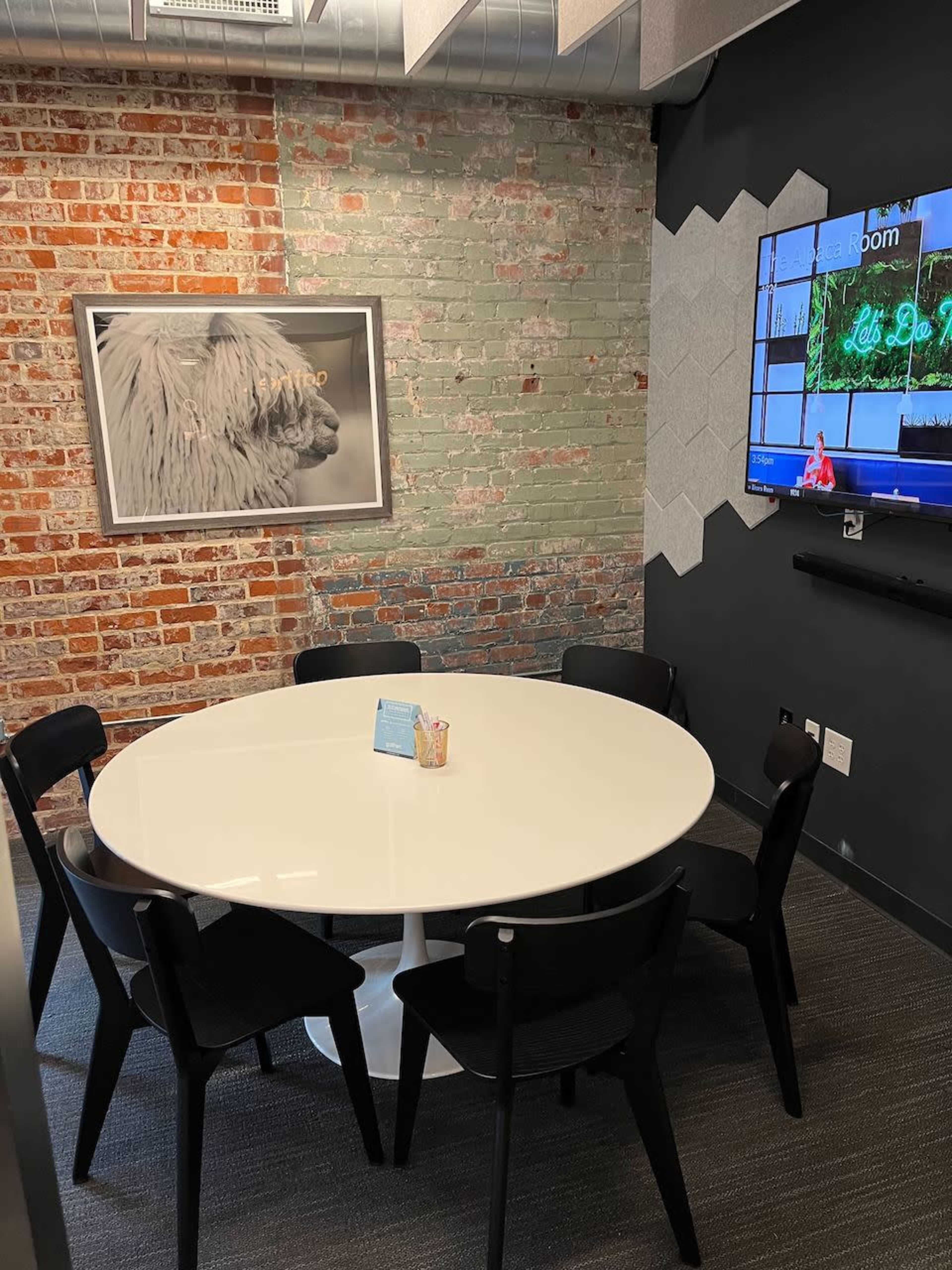 A conference room with a round white table surrounded by black chairs, a brick wall, and a television mounted on a gray wall.