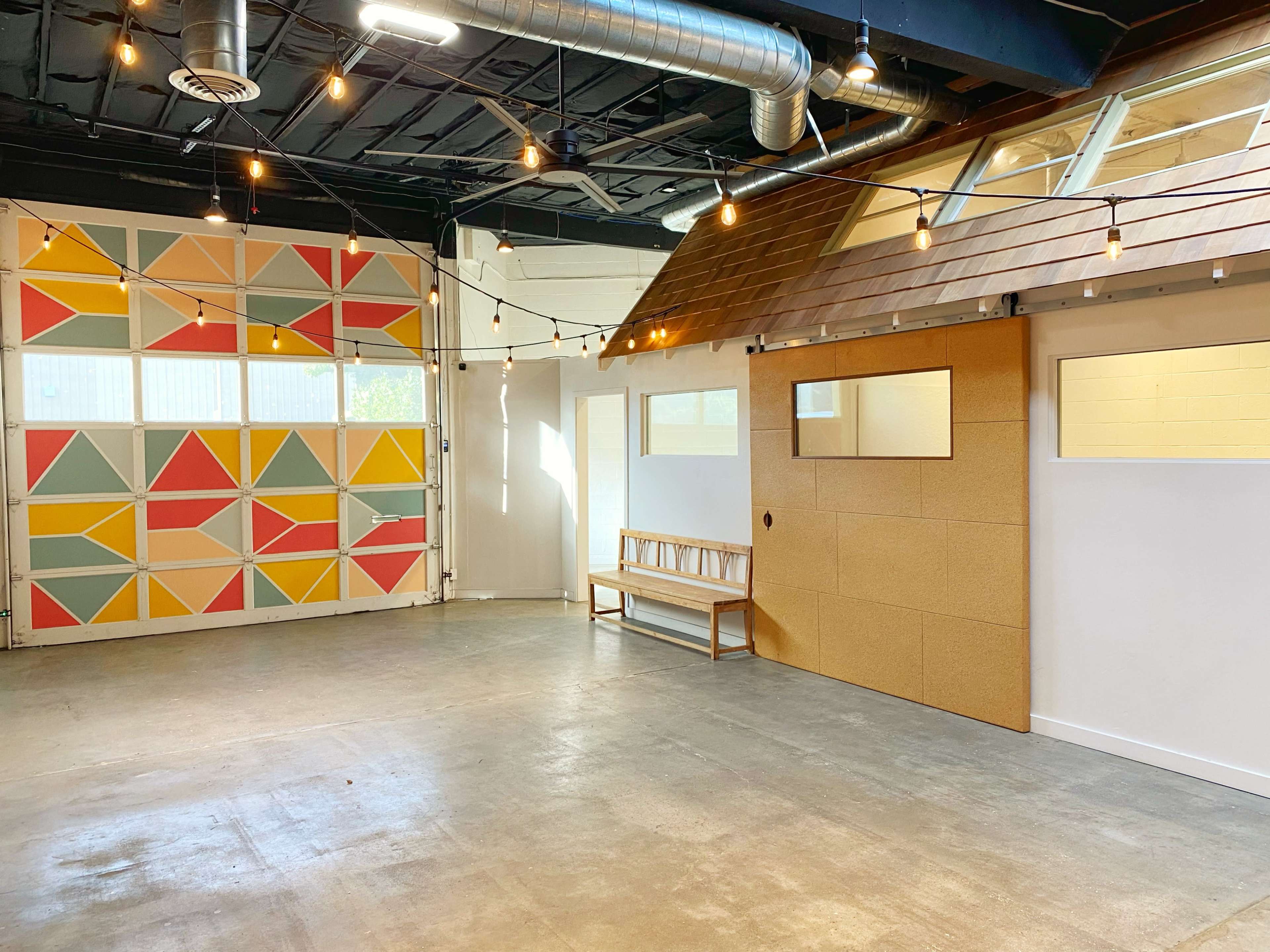 A spacious interior room with a colorful geometric mural on one wall, a wooden bench against another wall, and string lights hanging overhead.