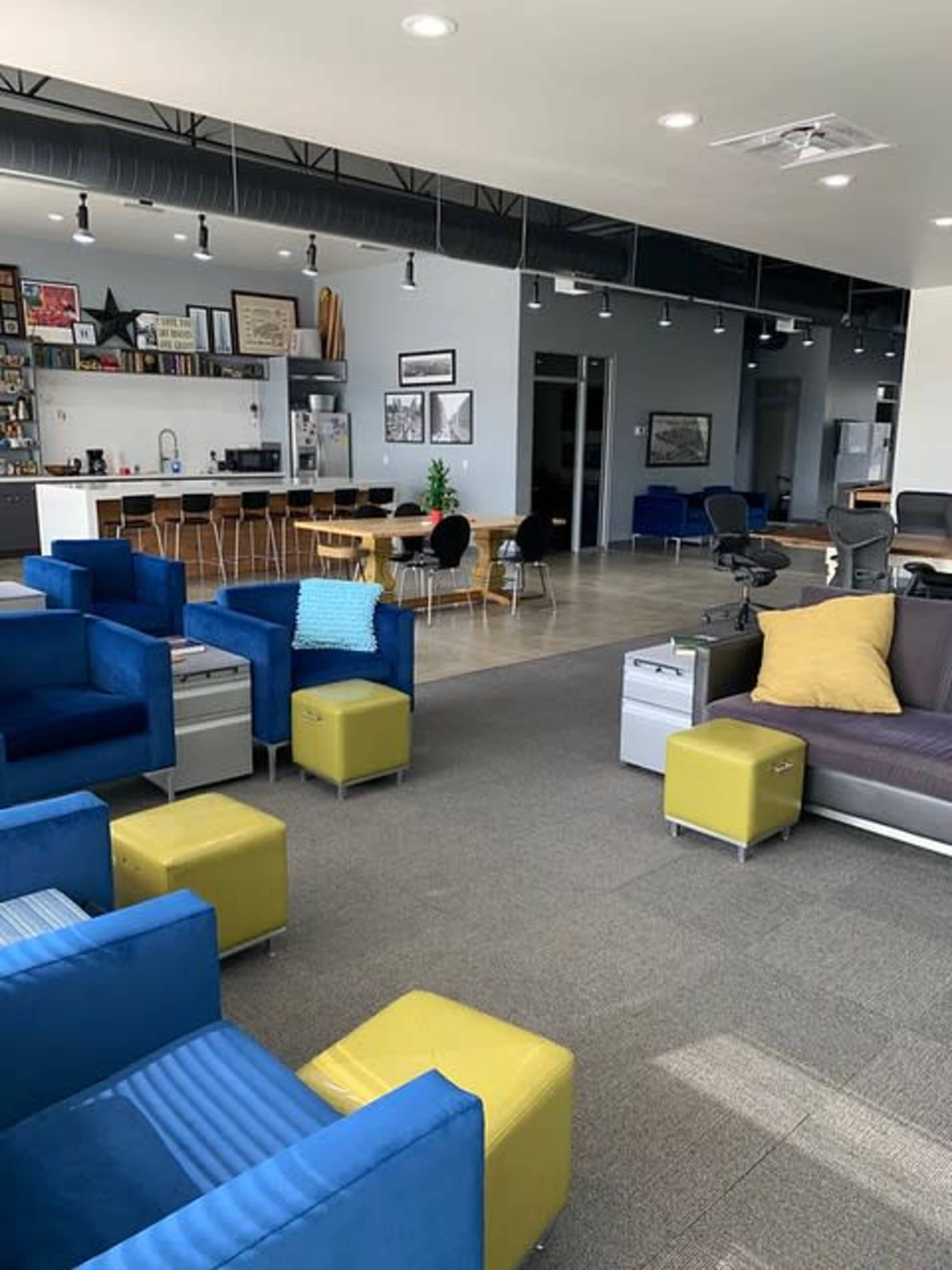 A modern office space with blue and yellow seating areas, a kitchen in the background, and a dining table with chairs.