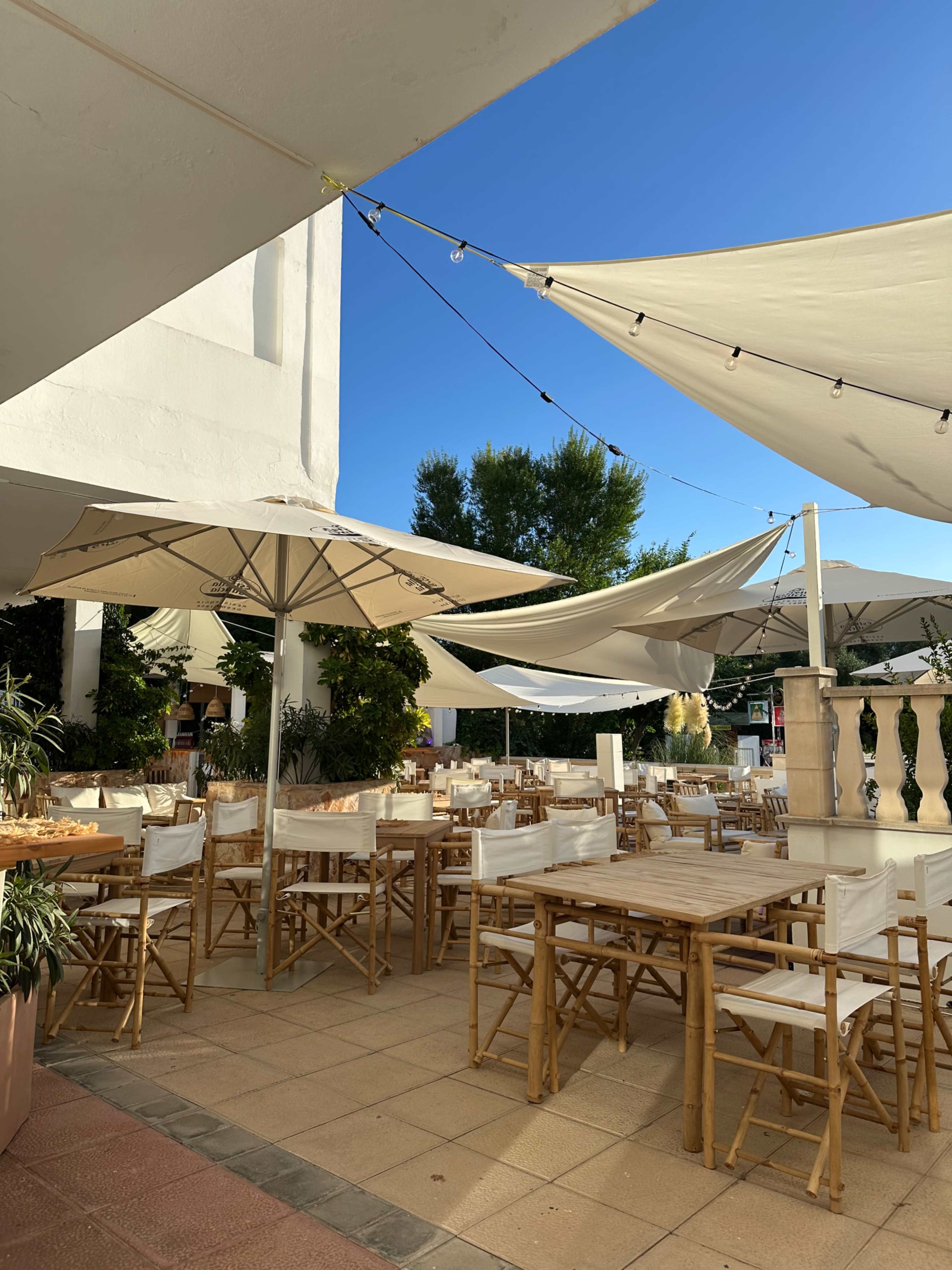 A dining area features bamboo tables and chairs under umbrellas, with a clear blue sky and shaded tents overhead.