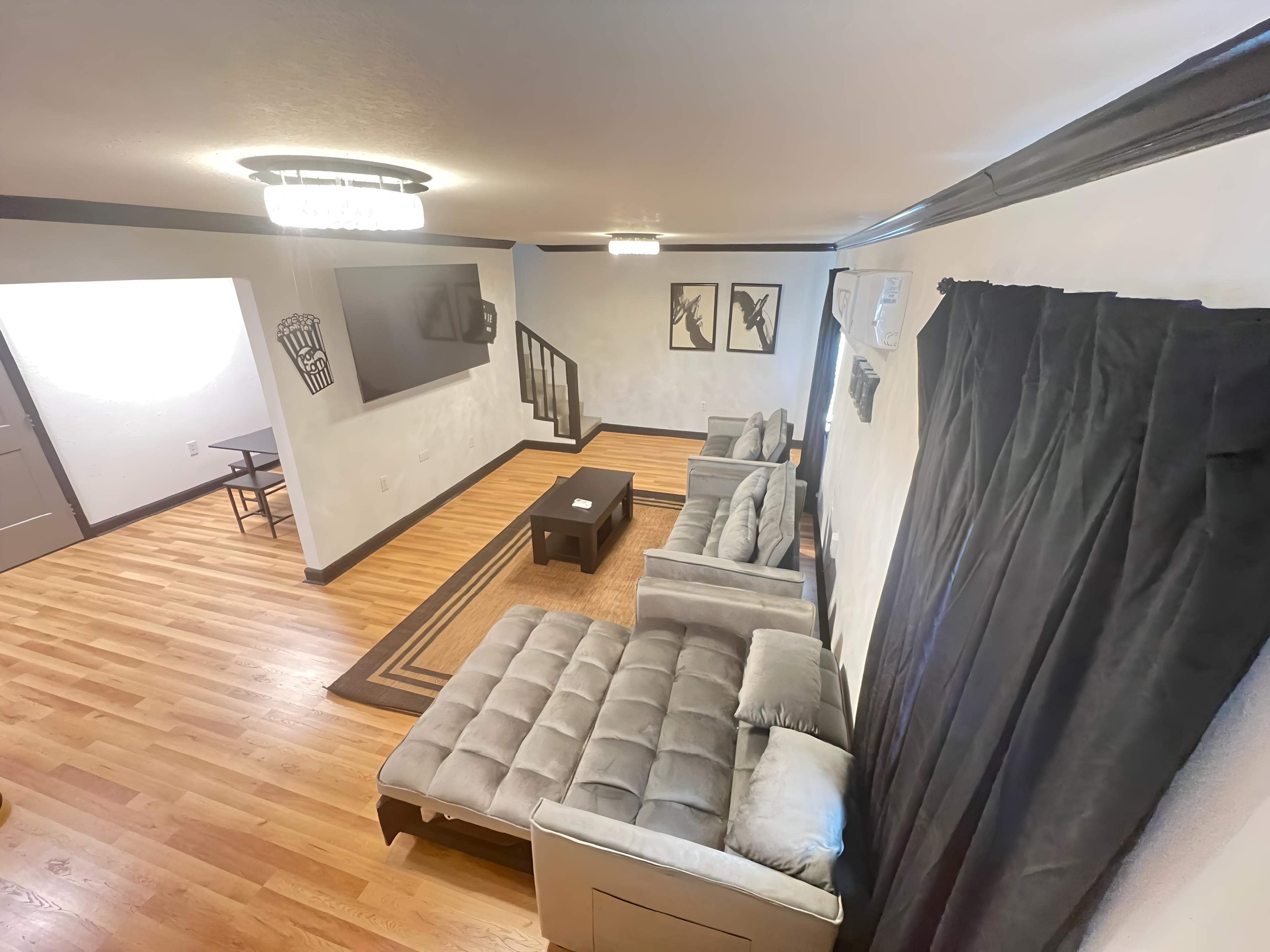 The image shows a living room featuring a sectional sofa, a coffee table, a mounted flat-screen TV, and a small dining area in the background.