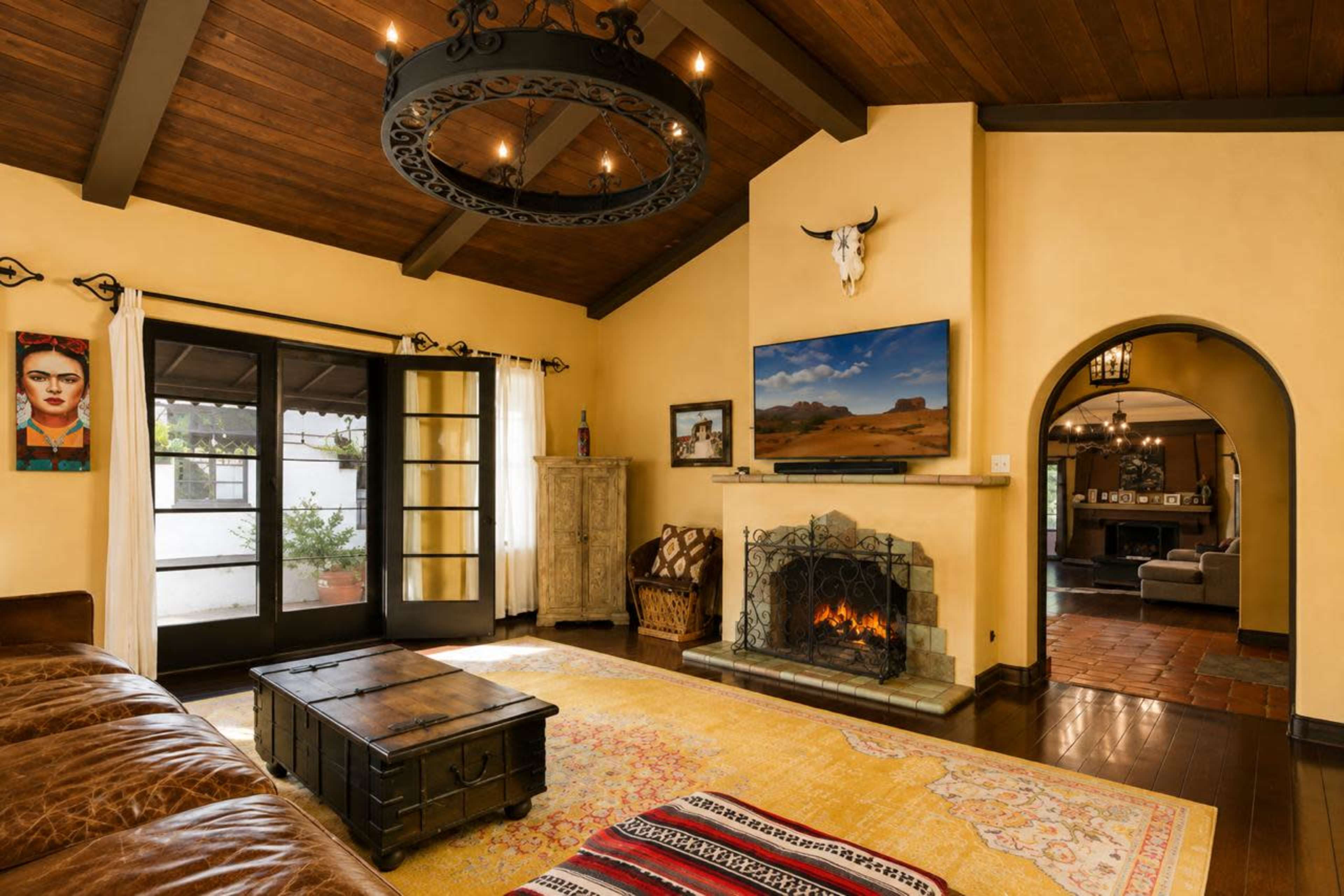 The image shows a cozy living room with a fireplace, a wooden ceiling, and a large TV mounted on the wall, decorated with a mix of modern and rustic elements.