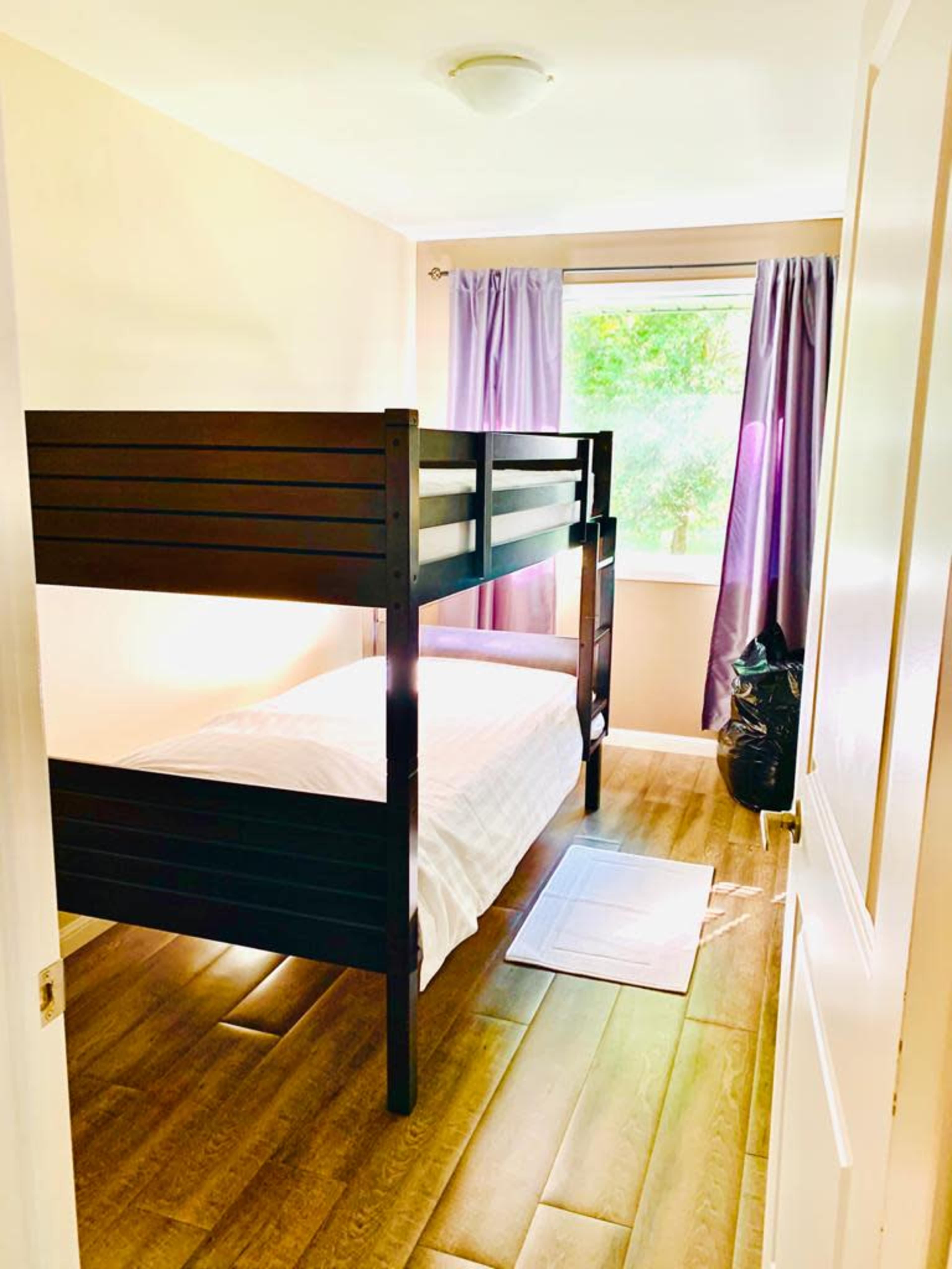 The image shows a small bedroom featuring a black bunk bed, a single bed on the bottom, a window with purple curtains, and a wooden floor.