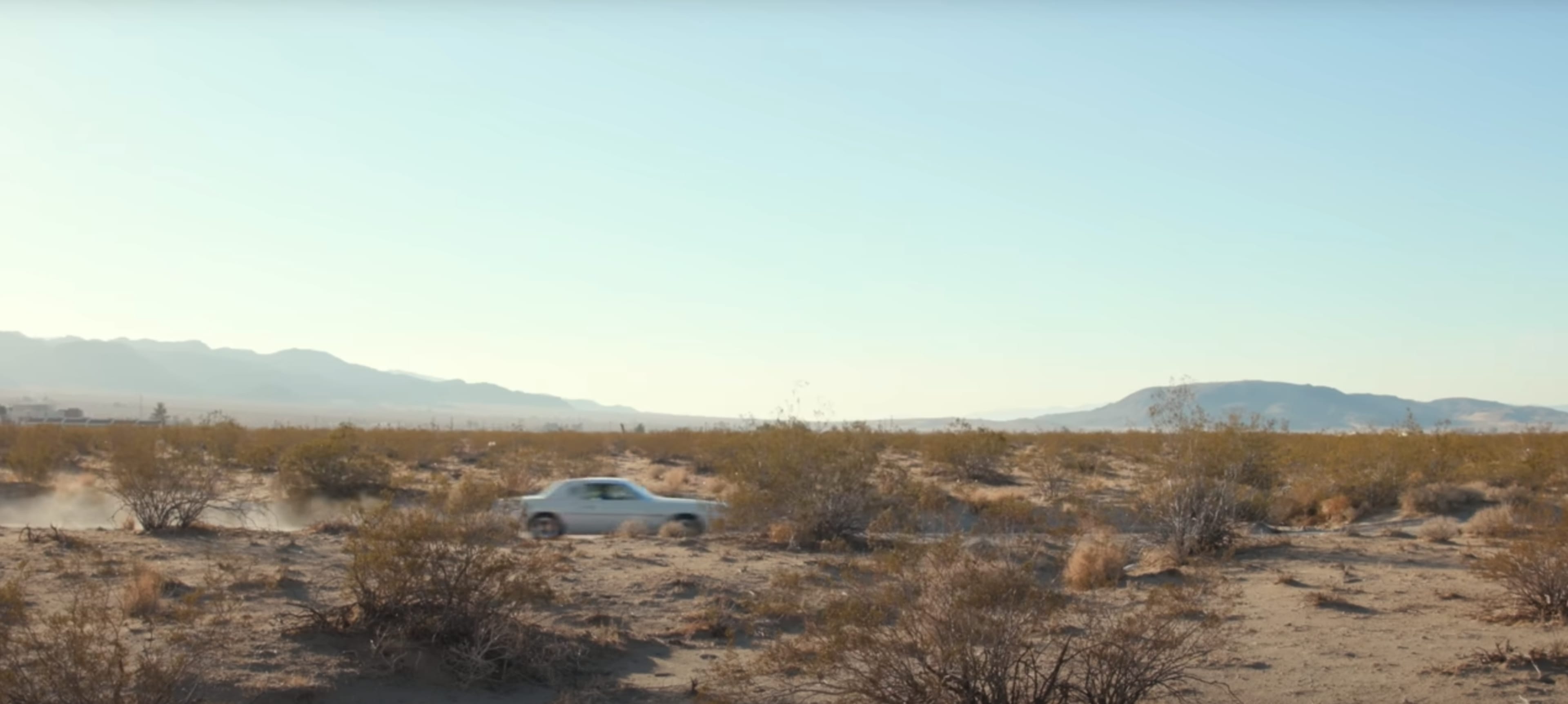 A white car drives through a sandy desert landscape with sparse vegetation and distant mountains.