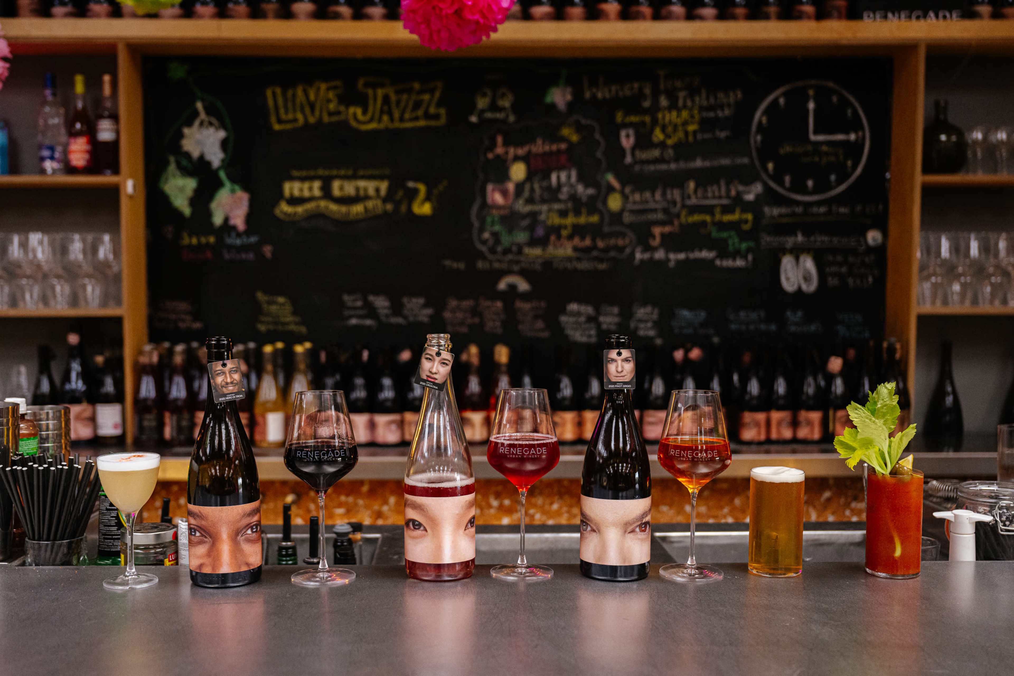 The image shows a bar countertop with various drinks and bottles, all featuring unique labels with faces, alongside a chalkboard displaying a "Live Jazz" event announcement and drink specials.