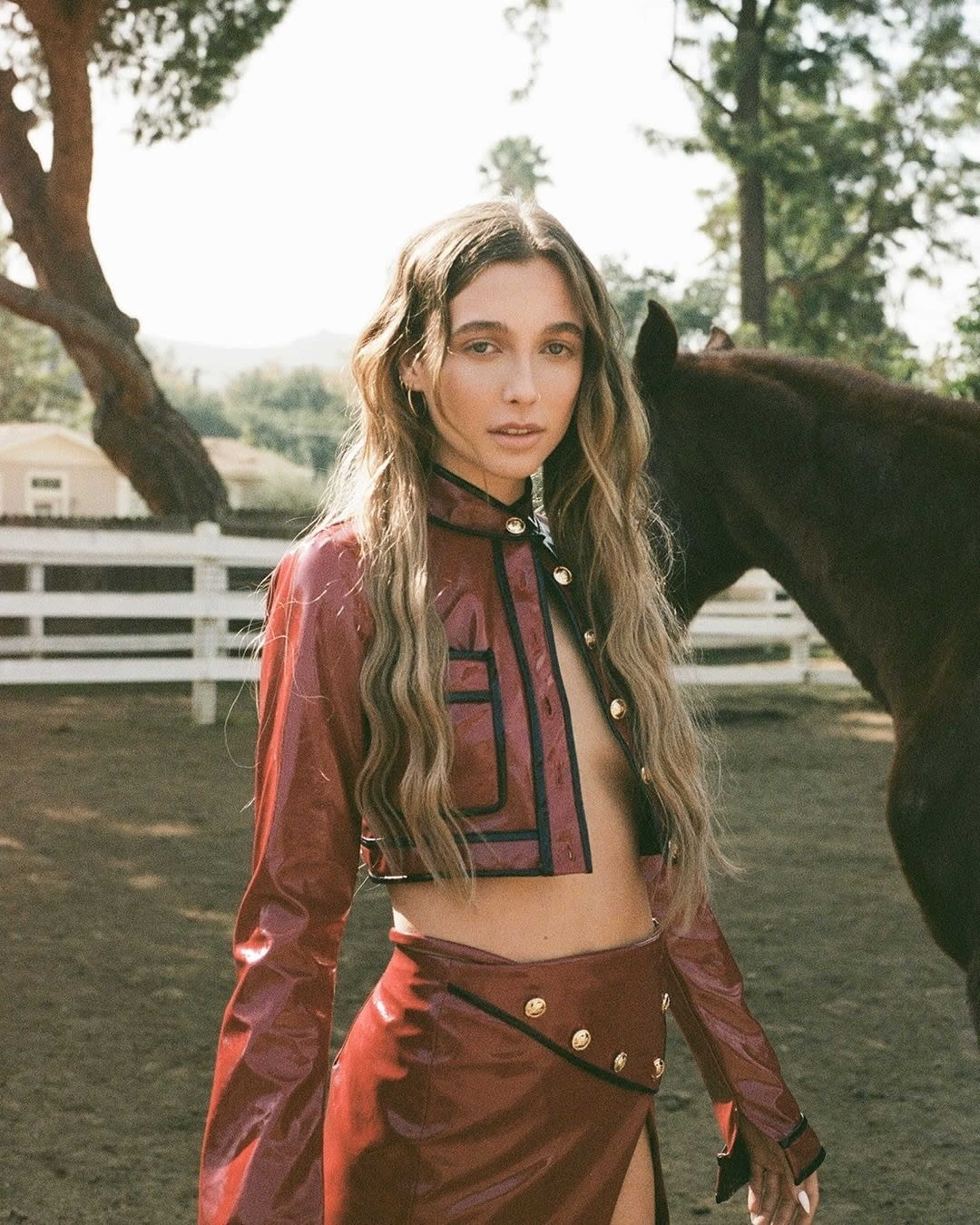 A person stands in a fenced area near a horse, wearing a red crop jacket and matching skirt, with trees in the background.